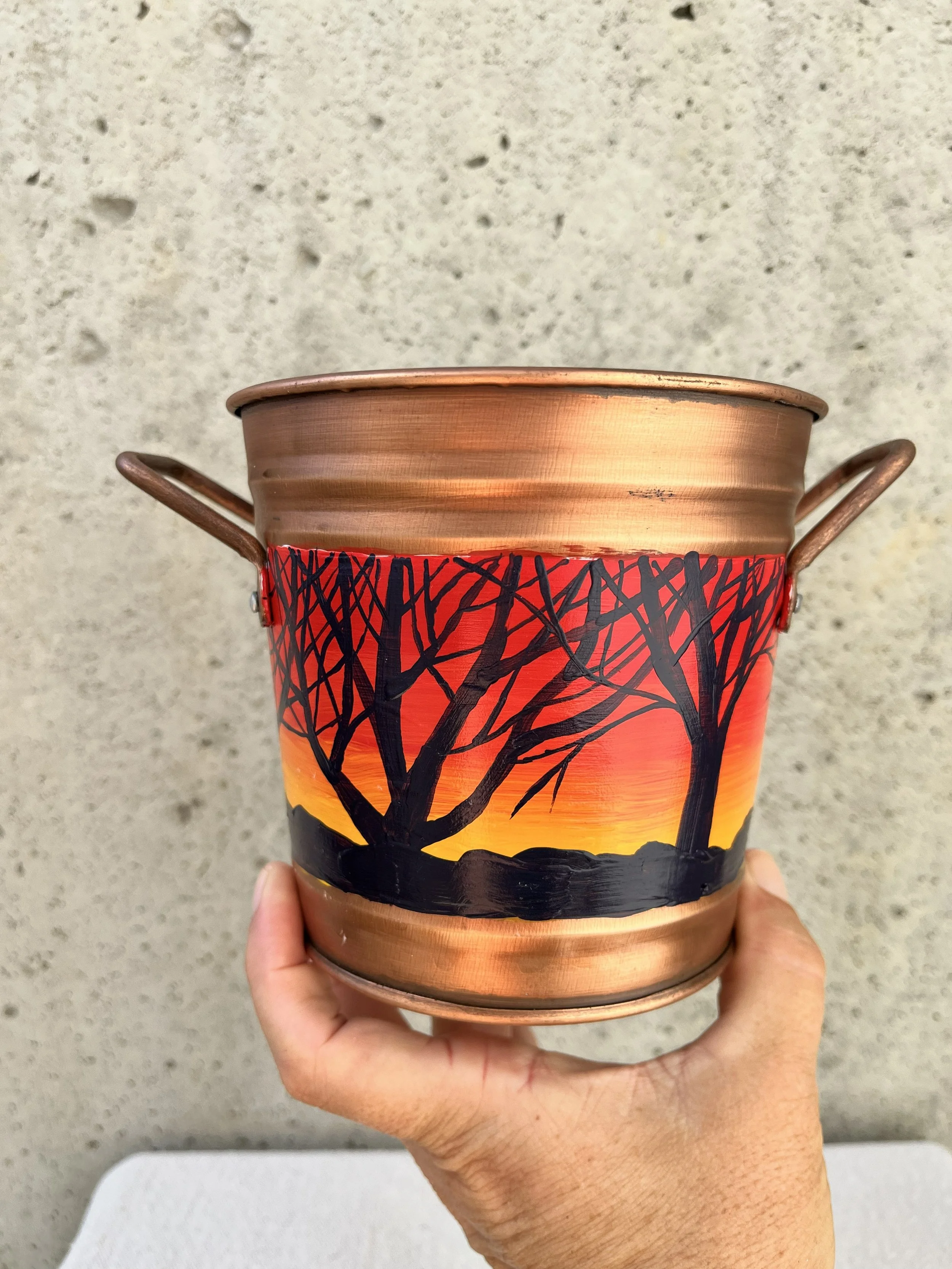 a handpainted landscape of bare branch trees silhouetted against a sunset. Painted on a copper color bucket.