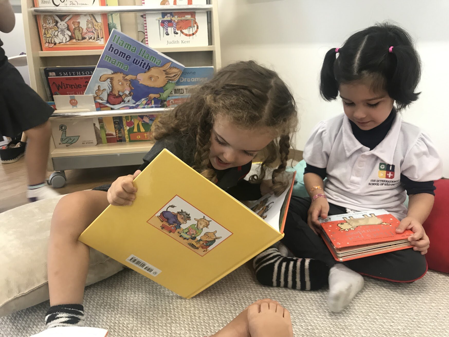 Two pre-school students in bilingual reading activity. Behind them is a shelf with other options of books in English.