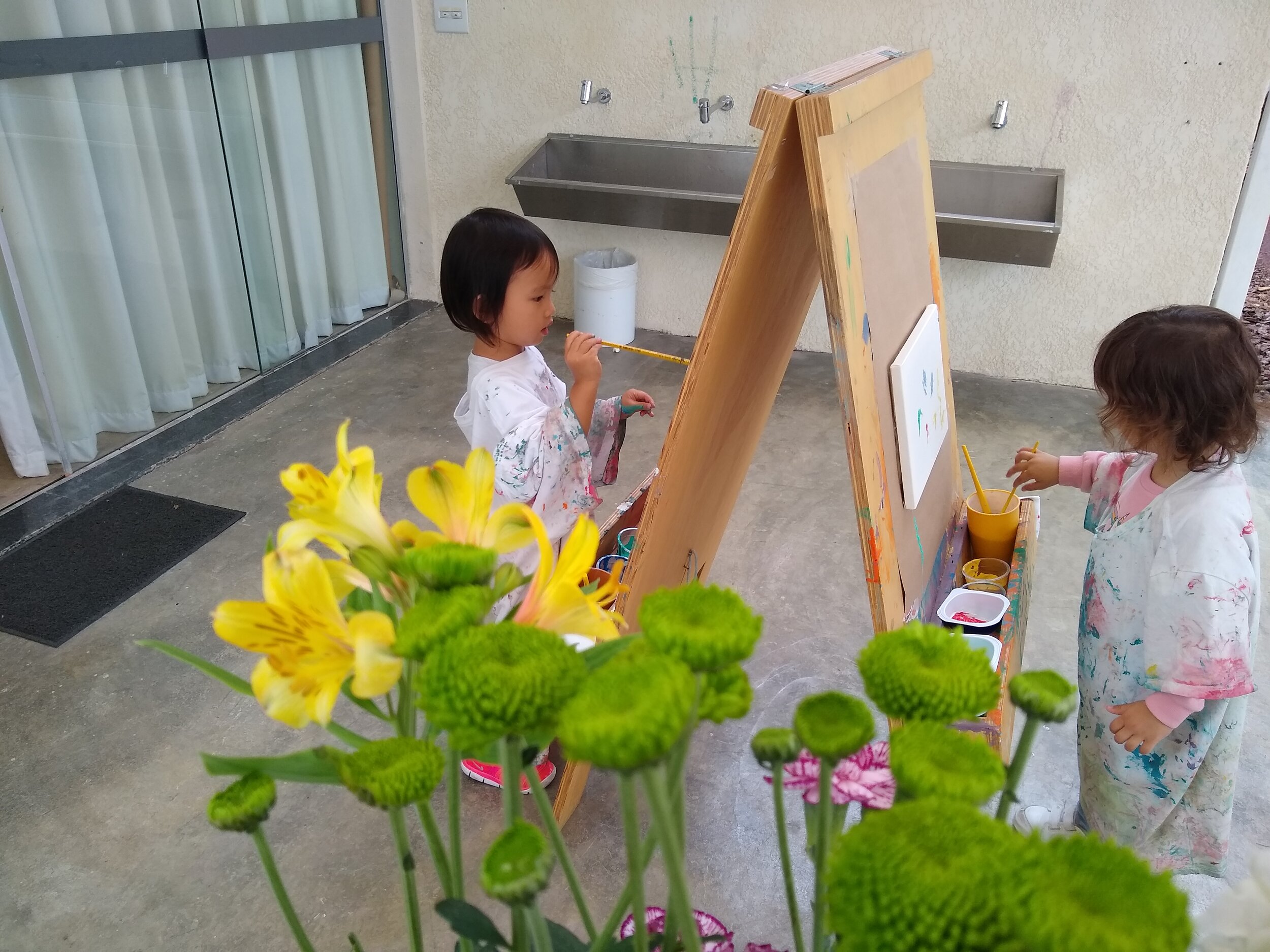 Two preschool students painting with an easel. They wear adult T-shirts, which cover the entire body, instead of the apron.