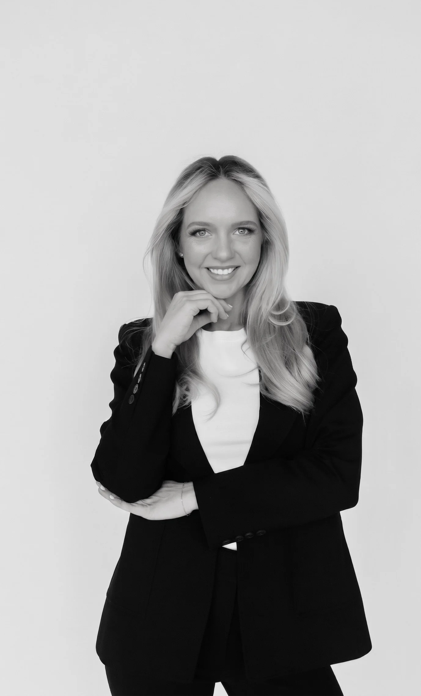 Black and white portrait of a smiling woman with long hair, wearing a blazer and white top, standing against a plain background.