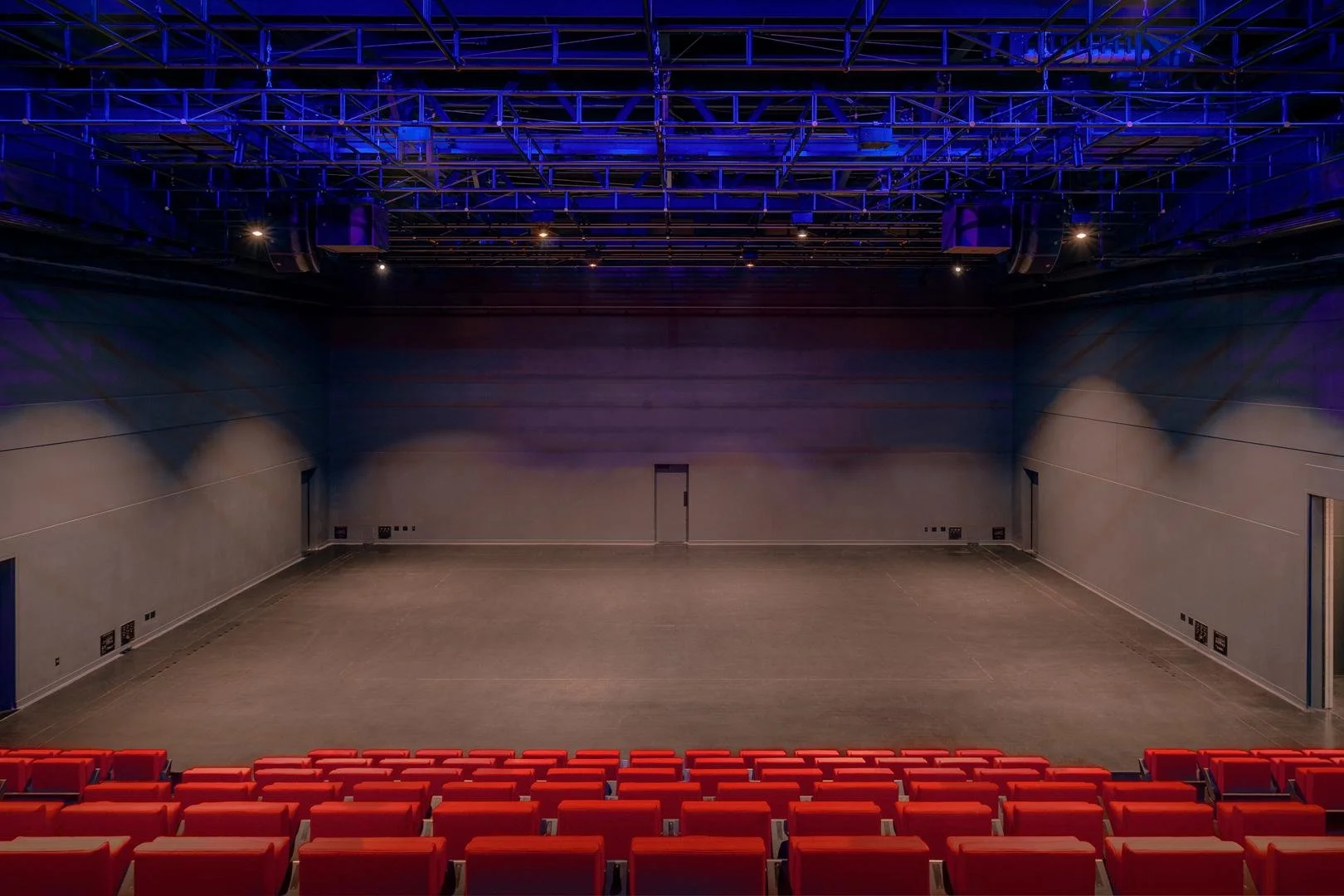 View looking down from the audience seating toward the empty flat stage floor of the black box theater illuminated by overhead blue lights.