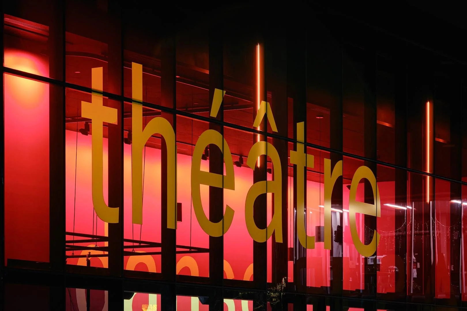 Gros plan sur le mot théâtre intégré à la façade vitrée du Grand Espace, montrant les jeux de reflets et la profondeur du foyer lumineux.