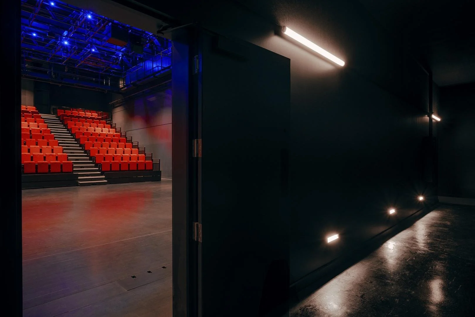 View from the dark acoustic vestibule looking through an open heavy black door into the bright theater hall with its red seating bank.
