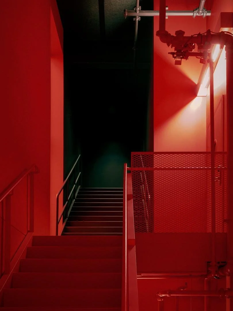 Striking monochrome red staircase featuring an expanded metal mesh guardrail and exposed industrial fire protection piping on the wall.