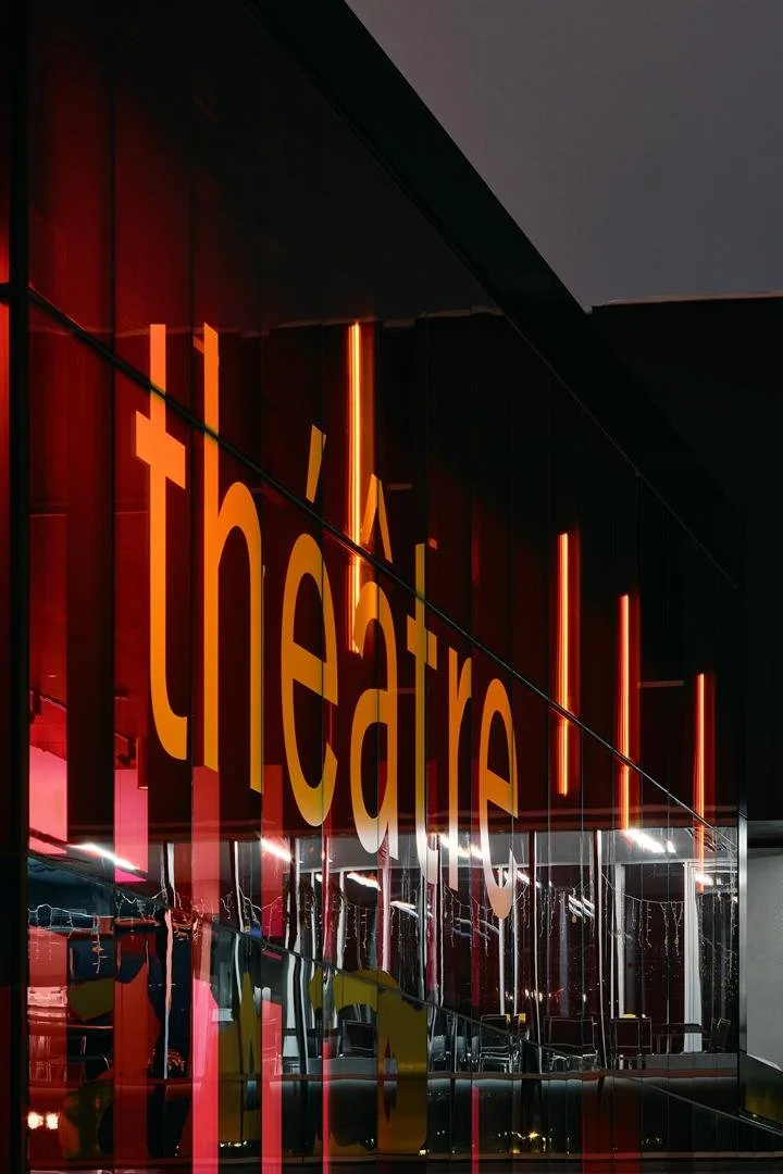 Close-up of the large yellow "théâtre" lettering applied to the glowing red glass facade of Le Grand-Espace at twilight.