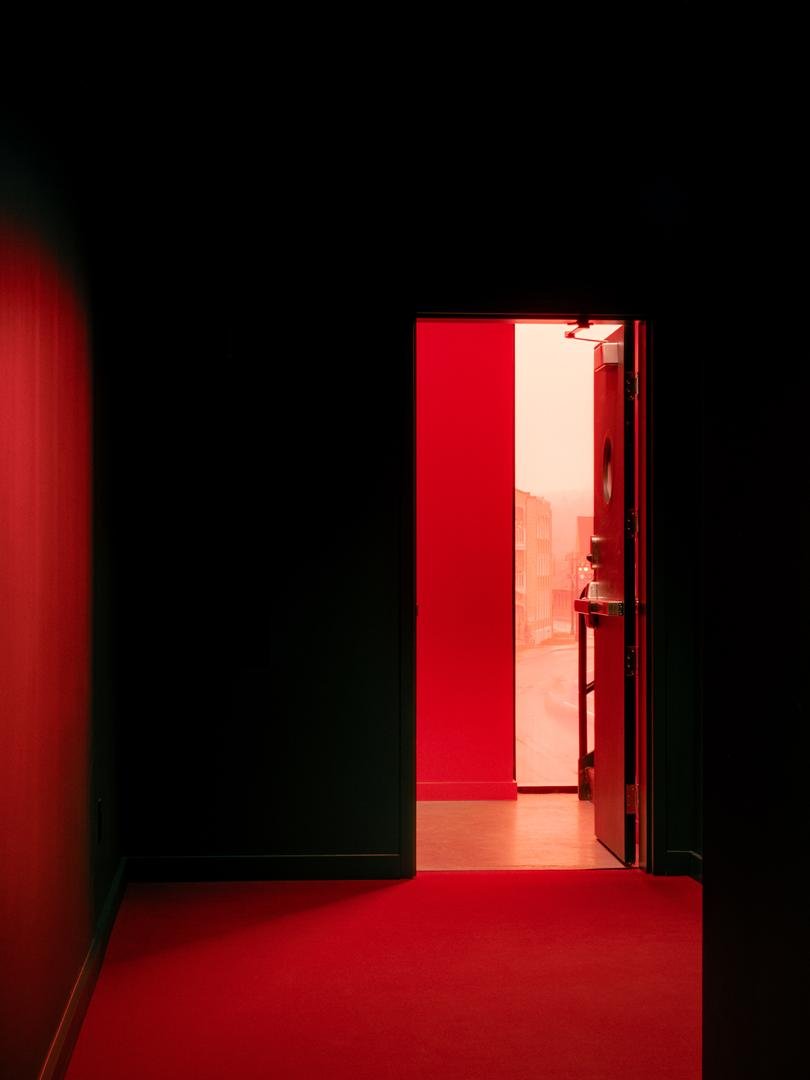 Contraste fort dans l'aménagement du Grand Espace : un couloir noir s'ouvre sur l'escalier avec une lumière naturelle teintée de rouge.