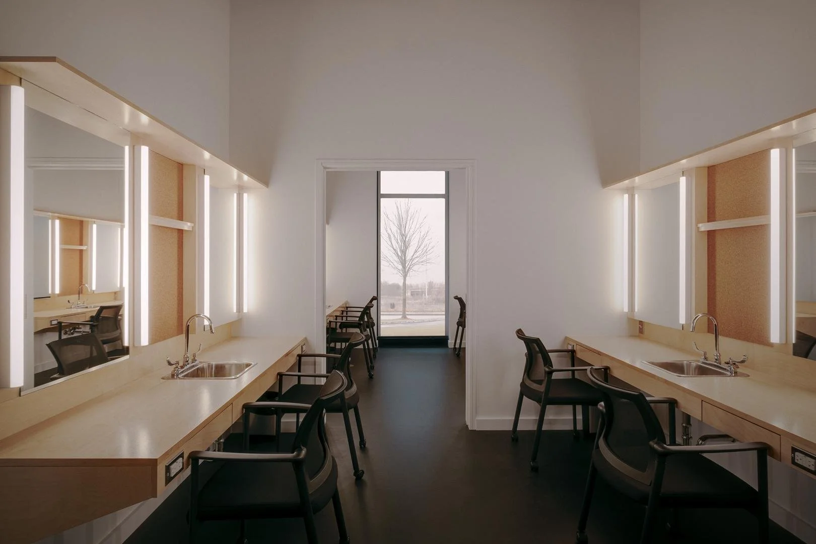 Bright modern artists dressing room featuring symmetrical light wood makeup stations with illuminated mirrors flanking a tall window.
