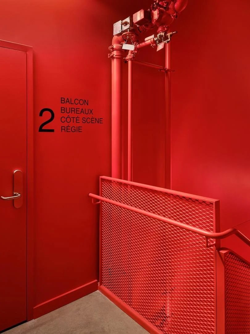 Cage d'escalier monochrome du Grand Espace entièrement peinte en rouge vif, incluant les portes, les garde-corps grillagés et la tuyauterie apparente, avec une signalétique noire pour le niveau 2.