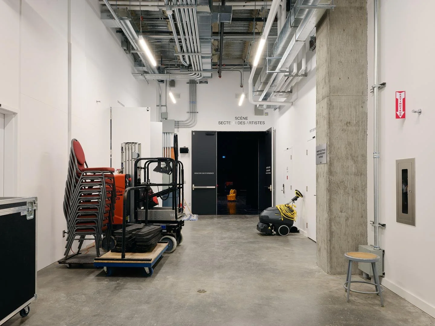 Backstage corridor in Le Grand-Espace showing exposed concrete column, stored equipment and double doors leading directly to the main stage.