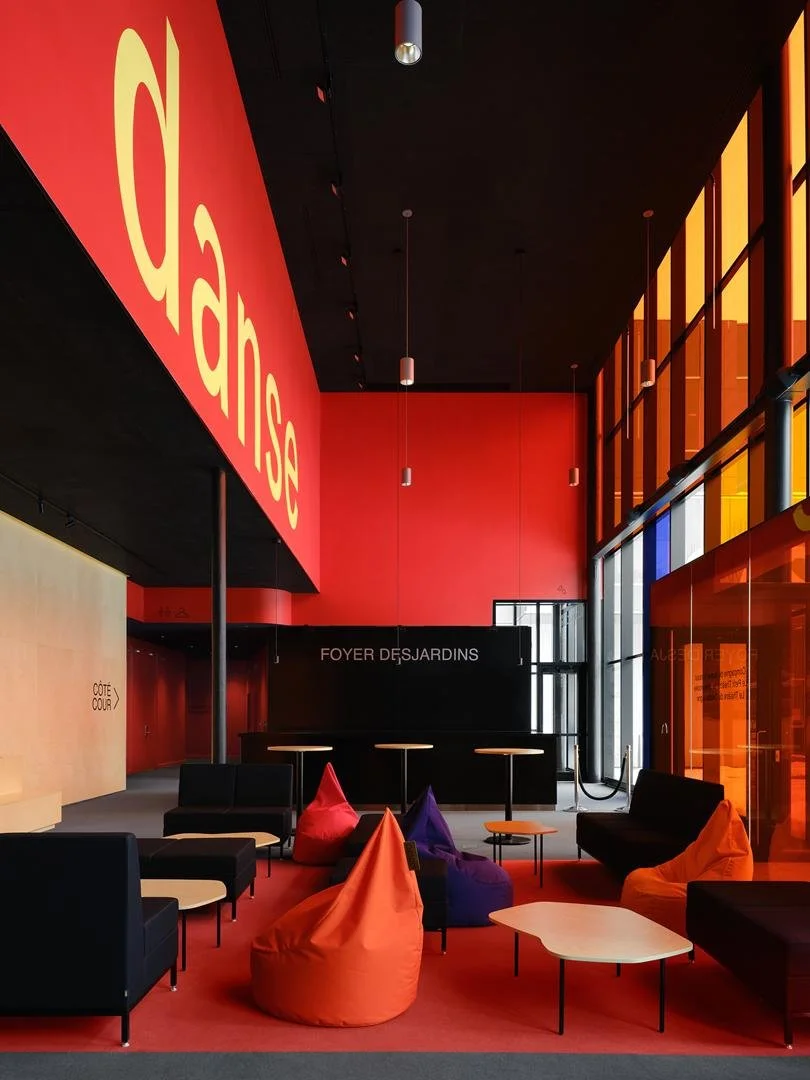 Espace d'accueil du Grand Espace avec un grand lettrage jaune épelant Danse sur un mur rouge vif, surplombant le Foyer Desjardins et ses sièges colorés.