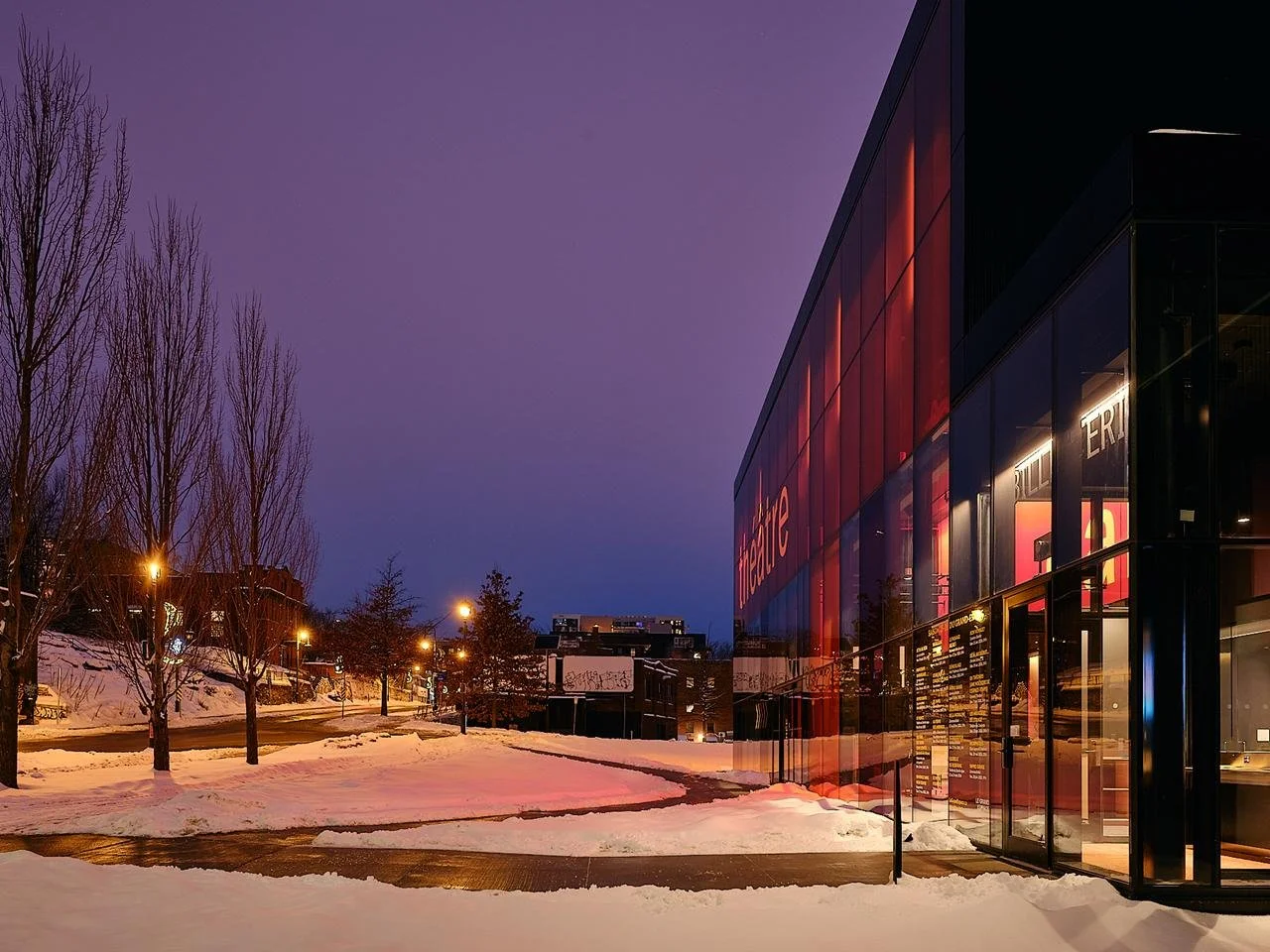 Evening perspective along the street, showing the illuminated red glass wall of Le Grand-Espace and its warm light spilling onto the winter snow.