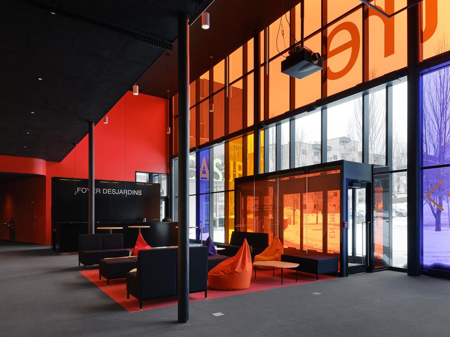 Vaste foyer vitré et lumineux du Grand Espace de Sherbrooke avec vitrages avec pellicules de couleurs rouges, orangés et mauves, aménagé comme une scène urbaine accueillante.