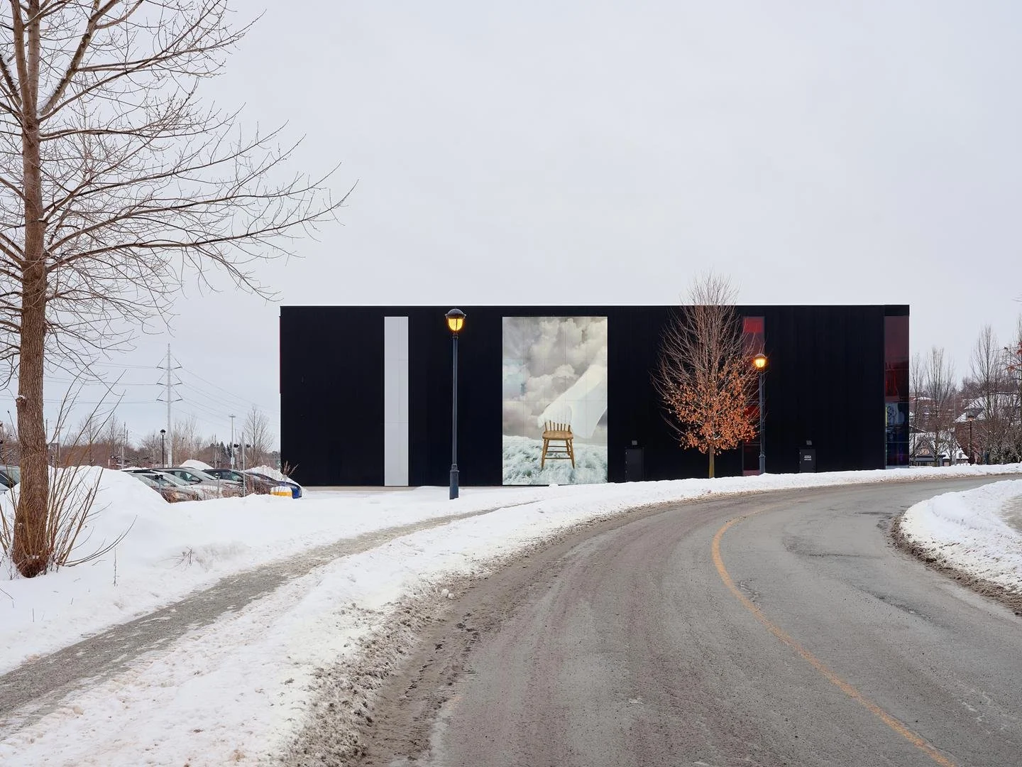 The black corrugated metal lateral facade of Le Grand-Espace, acting as a canvas for a large, integrated public artwork depicting a chair floating in clouds.