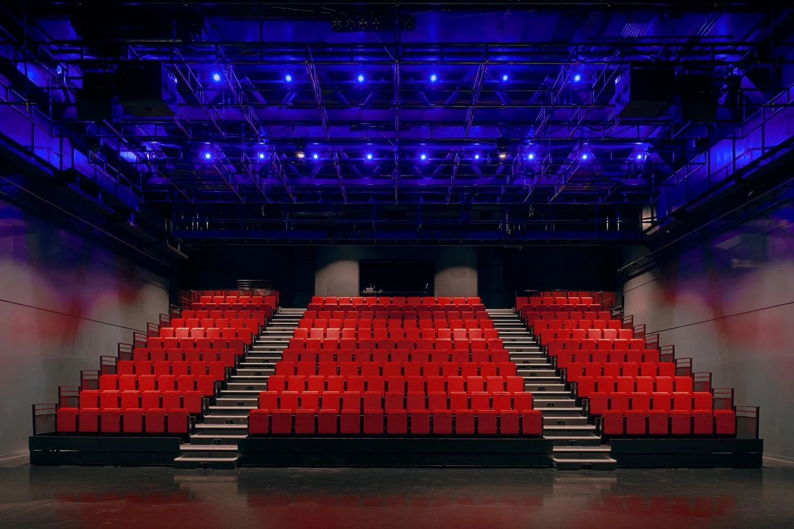 Symmetrical frontal view of the deployed red motorized seating bleacher inside the black box theater under a dramatic blue lighting grid.