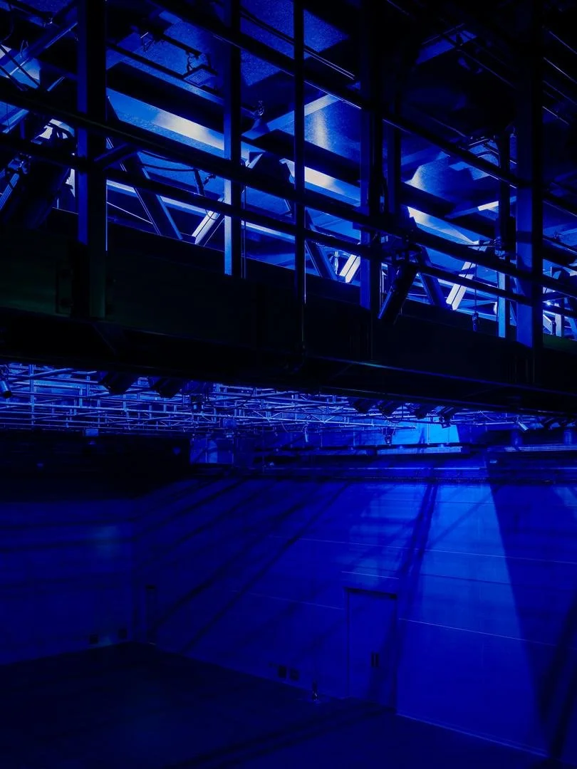 Upward view of the suspended steel technical catwalk casting long dramatic shadows against the theater walls under deep blue stage lights.