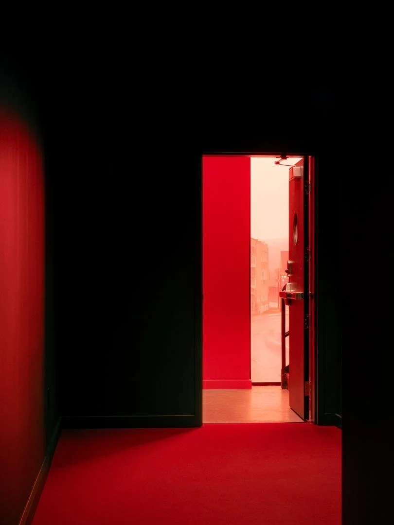 Open exit door in a dark corridor revealing a brightly illuminated vivid red interior stairway and a glimpse of the street outside.