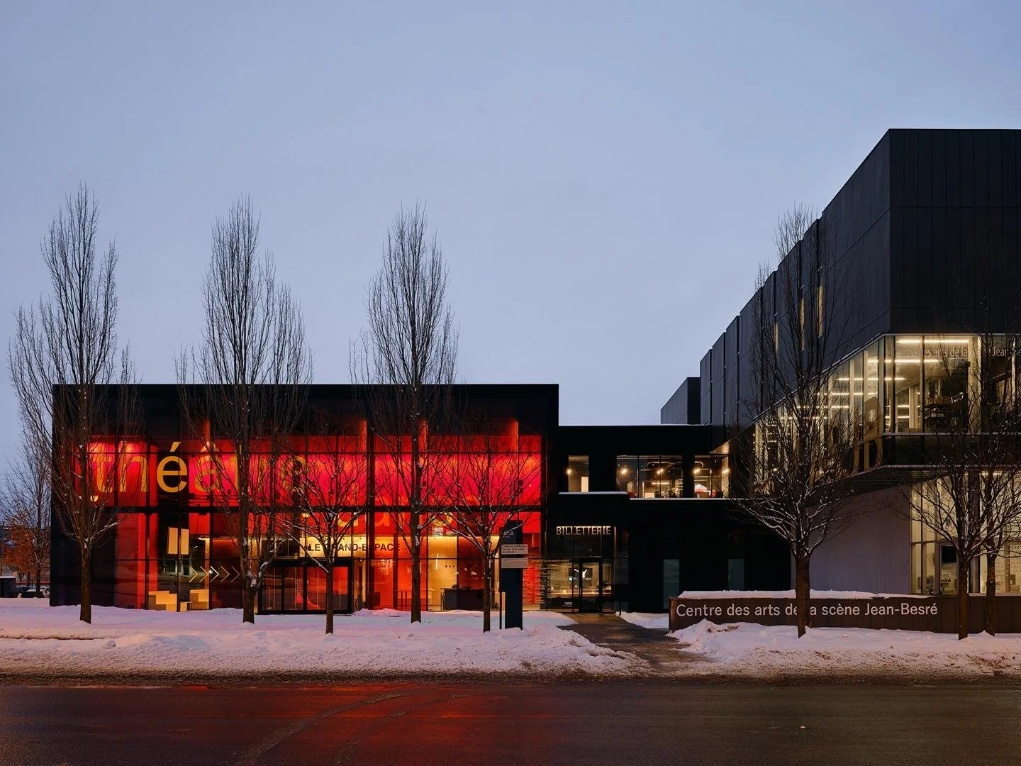 Nighttime view of Le Grand-Espace theater radiating red light from its glass facade, connected to the darker volume of the CASJB theatrical production facility.