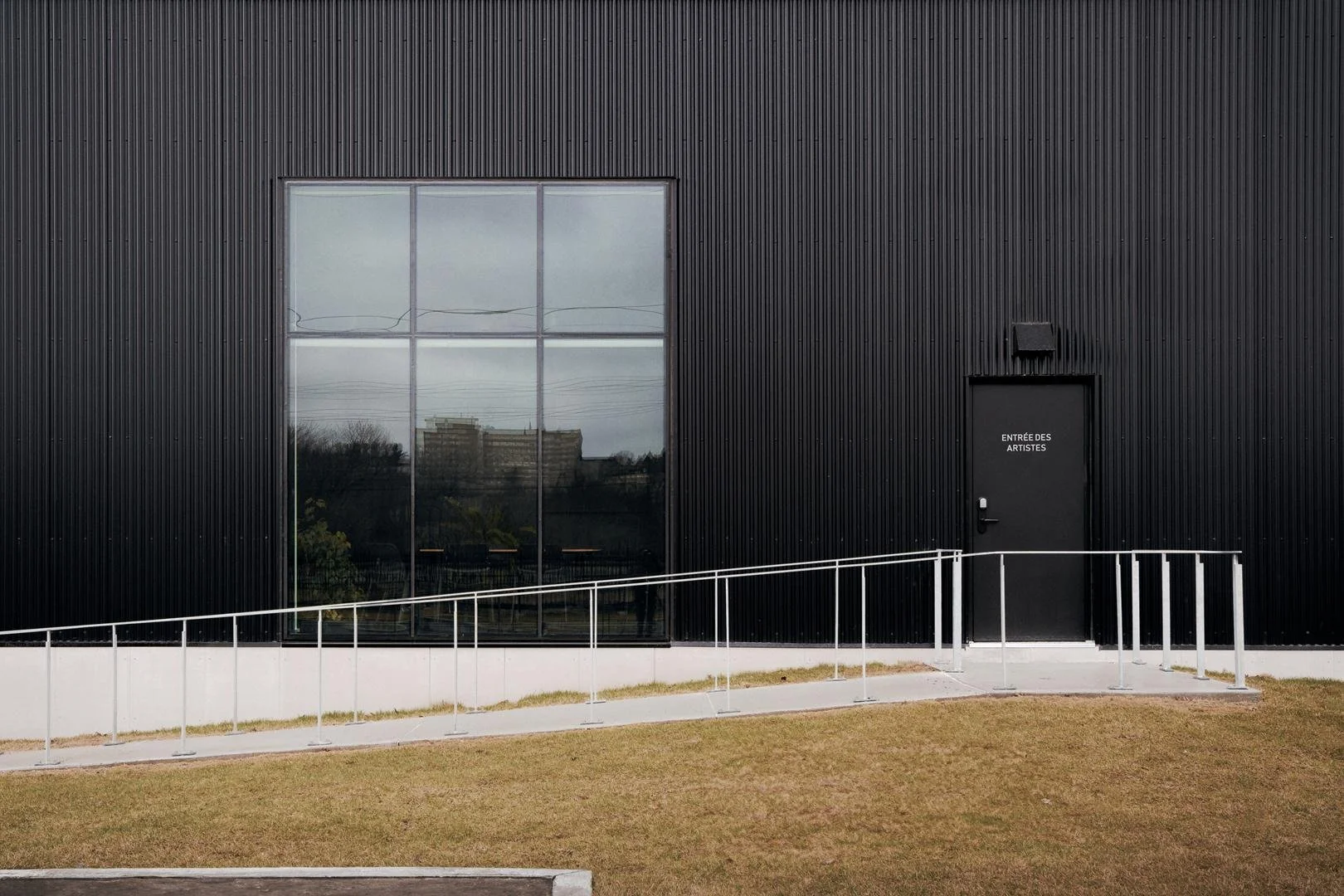 Minimalist black stage door labeled "Entrée des artistes" next to the large square window of the green room reflecting the city skyline on the black corrugated metal facade.