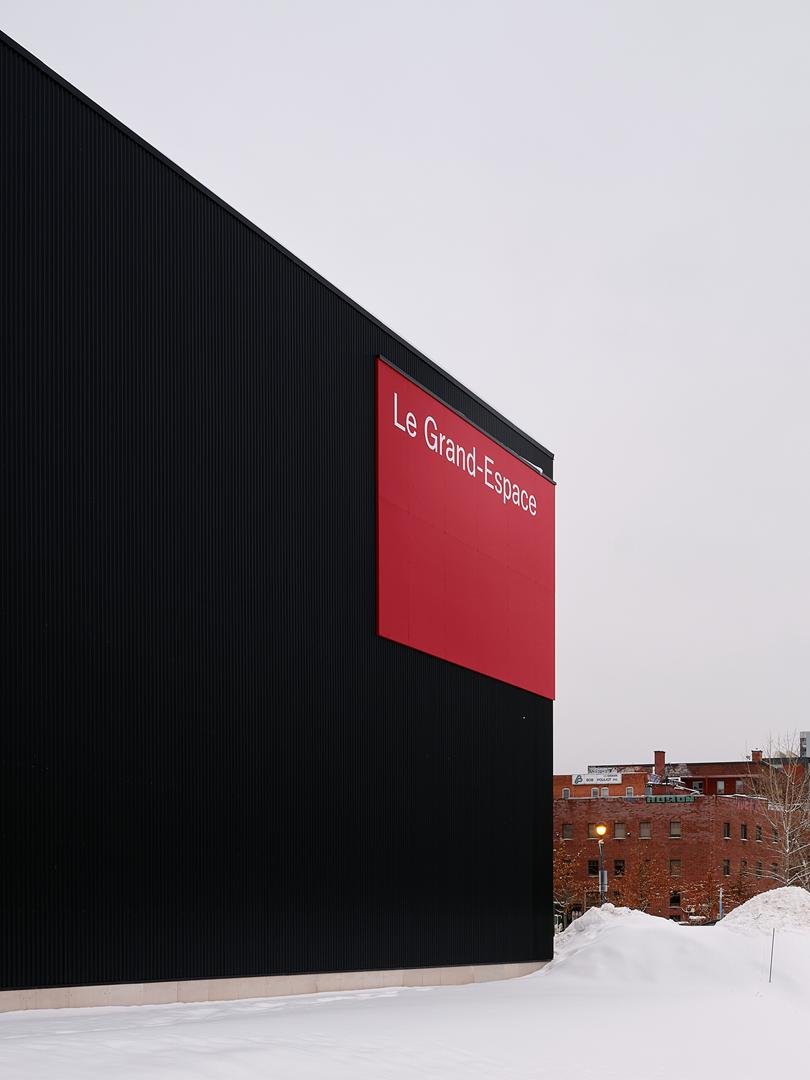 Gros plan sur la signalétique extérieure du Grand Espace : un large panneau rouge avec lettrage blanc fixé sur le revêtement métallique noir du bâtiment.