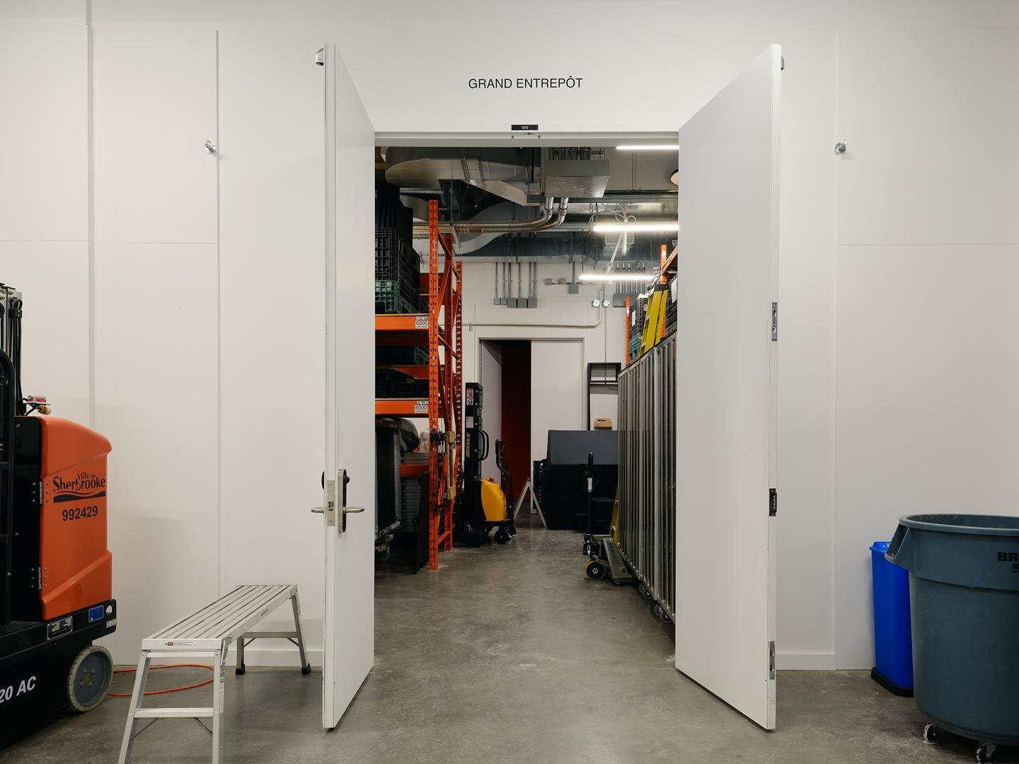 Grandes portes doubles blanches ouvertes sur le Grand Entrepôt du complexe, révélant un espace de rangement haut de plafond avec tablettes industrielles.