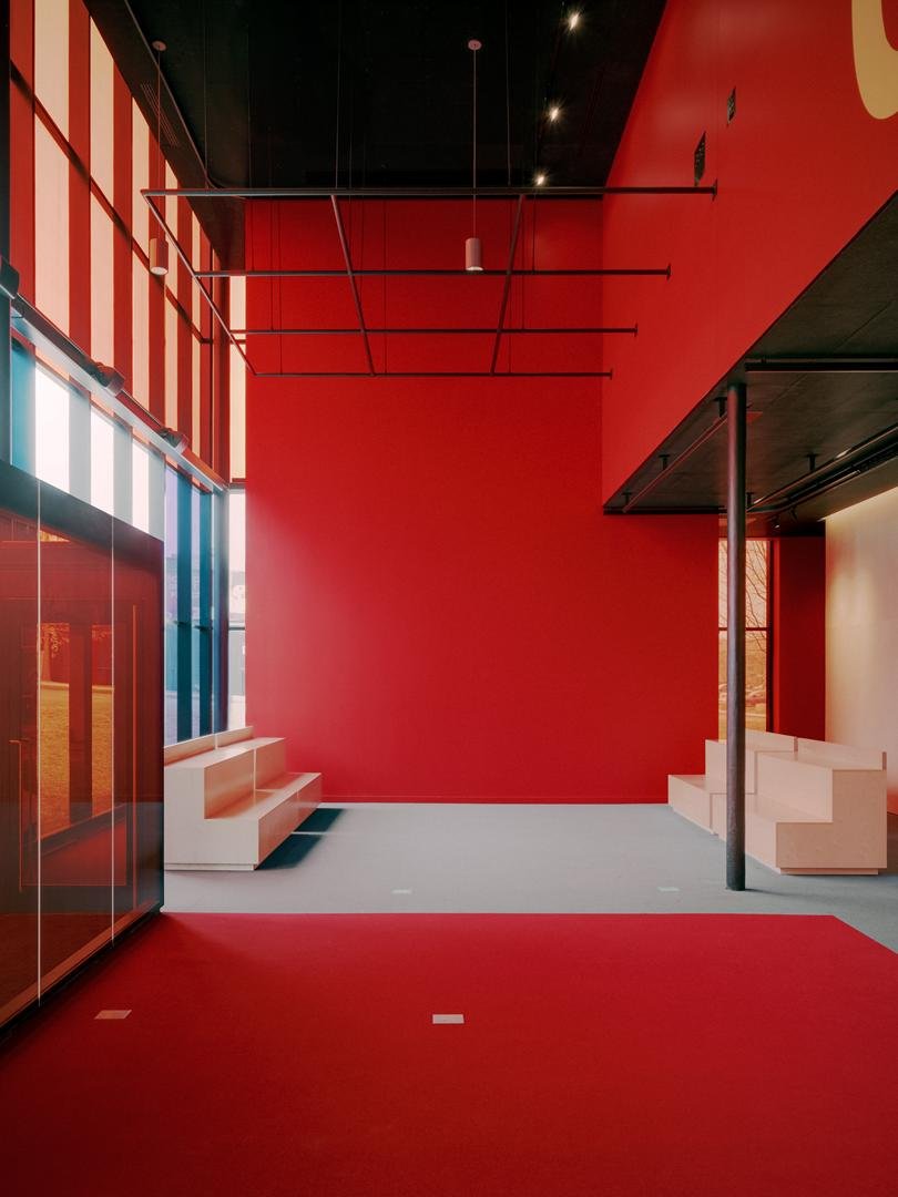 Foyer monumental du Grand Espace avec un mur monochrome rouge, un plafond acoustique noir et de petits gradins en bois clair pour le jeune public.
