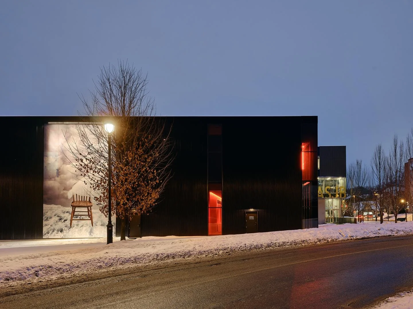 The lateral facade of Le Grand-Espace at twilight, highlighting the illuminated integrated public artwork of a chair against a cloudy sky.
