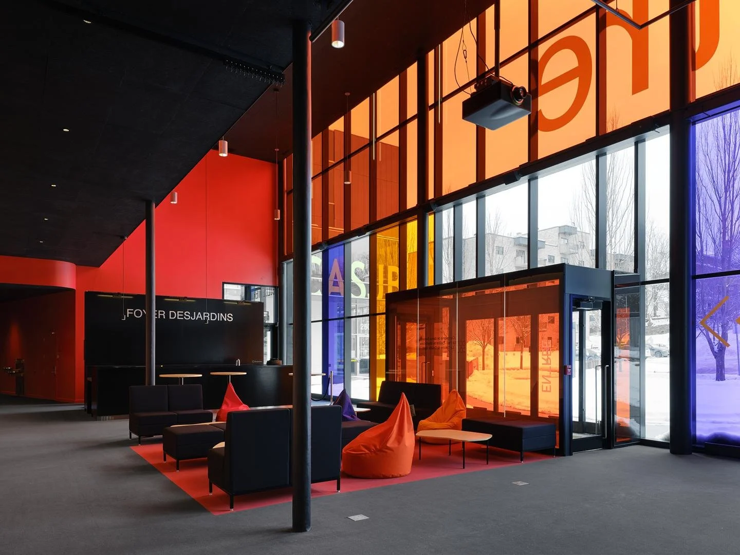 Interior view from the Le Grand-Espace foyer, showing colorful tinted glass panels in red, orange, and purple framing the snowy urban scene of Sherbrooke outside.