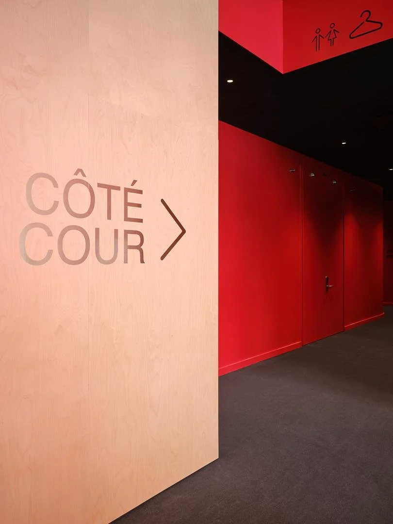 Mur en contreplaqué clair avec lettrage Côté Cour adjacent à un couloir rouge menant à la salle et au vestiaire, démontrant le design signalétique du Grand Espace.