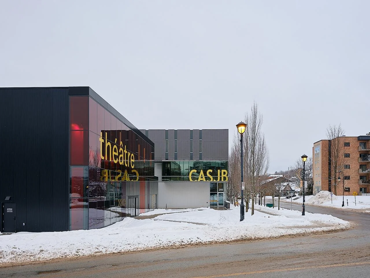 Daytime exterior view illustrating the complementary volumes of the red glass Le Grand-Espace theater and the attached CASJB production center in a snowy urban setting.