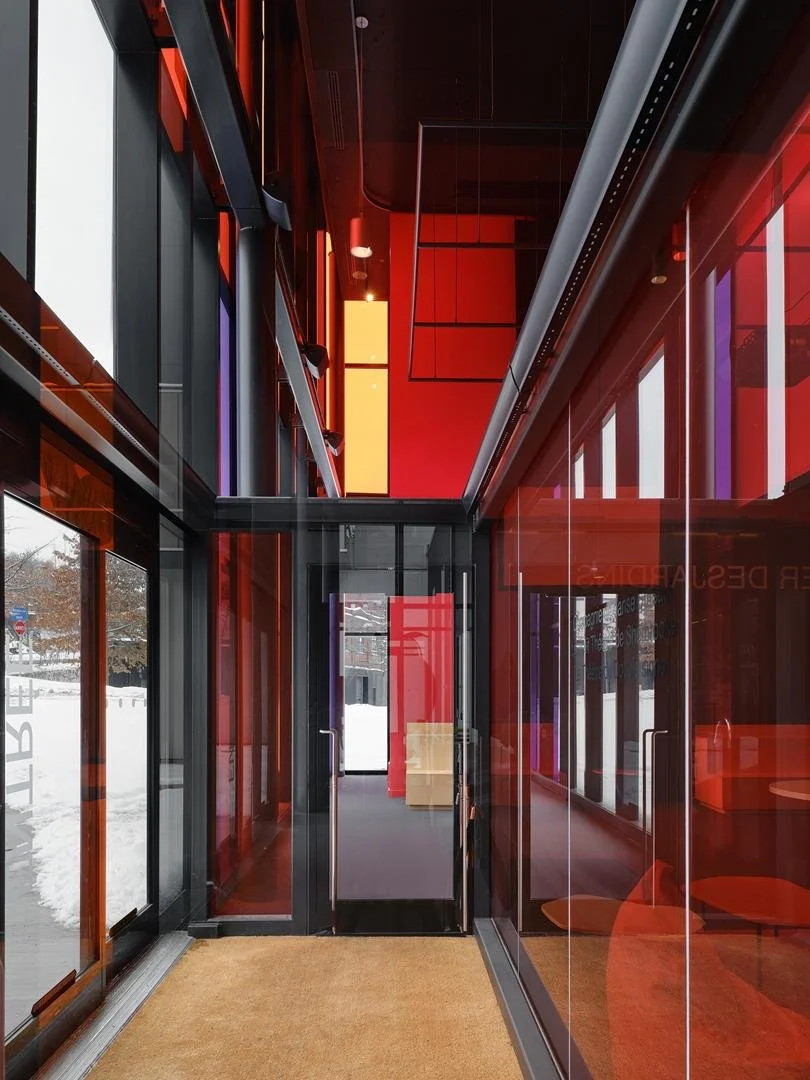 Vestibule lumineux donnant dans le foyer du Grand Espace, traversé par la lumière naturelle filtrée par des vitrages teintés de rouge, orange et mauve.