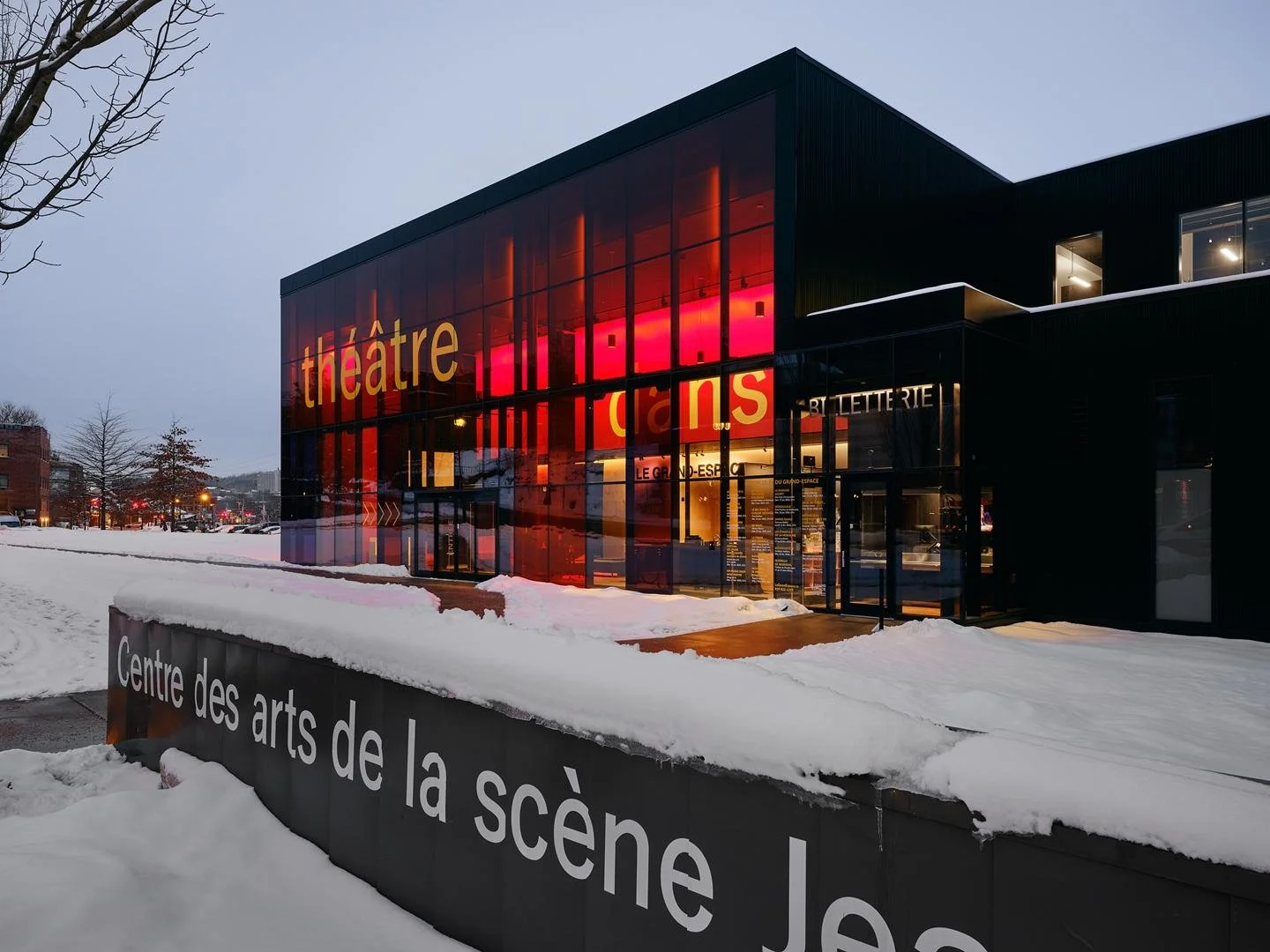 Vue au crépuscule du Grand Espace avec sa façade vitrée rougeoyante illuminée de l'intérieur, avec le muret d'entrée du Centre des arts de la scène Jean-Besré en avant plan.