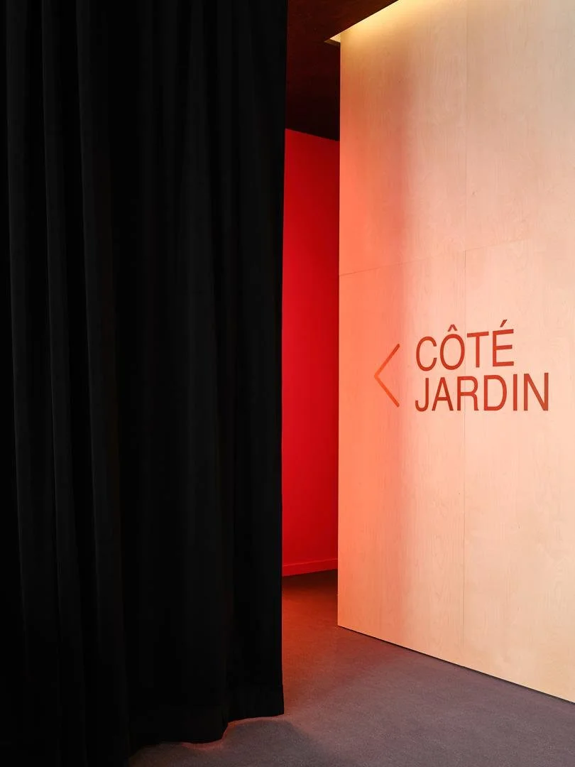 Signalétique intérieure du Grand Espace indiquant Côté Jardin avec un lettrage doré sur un mur en contreplaqué clair, derrière un rideau de velours noir.
