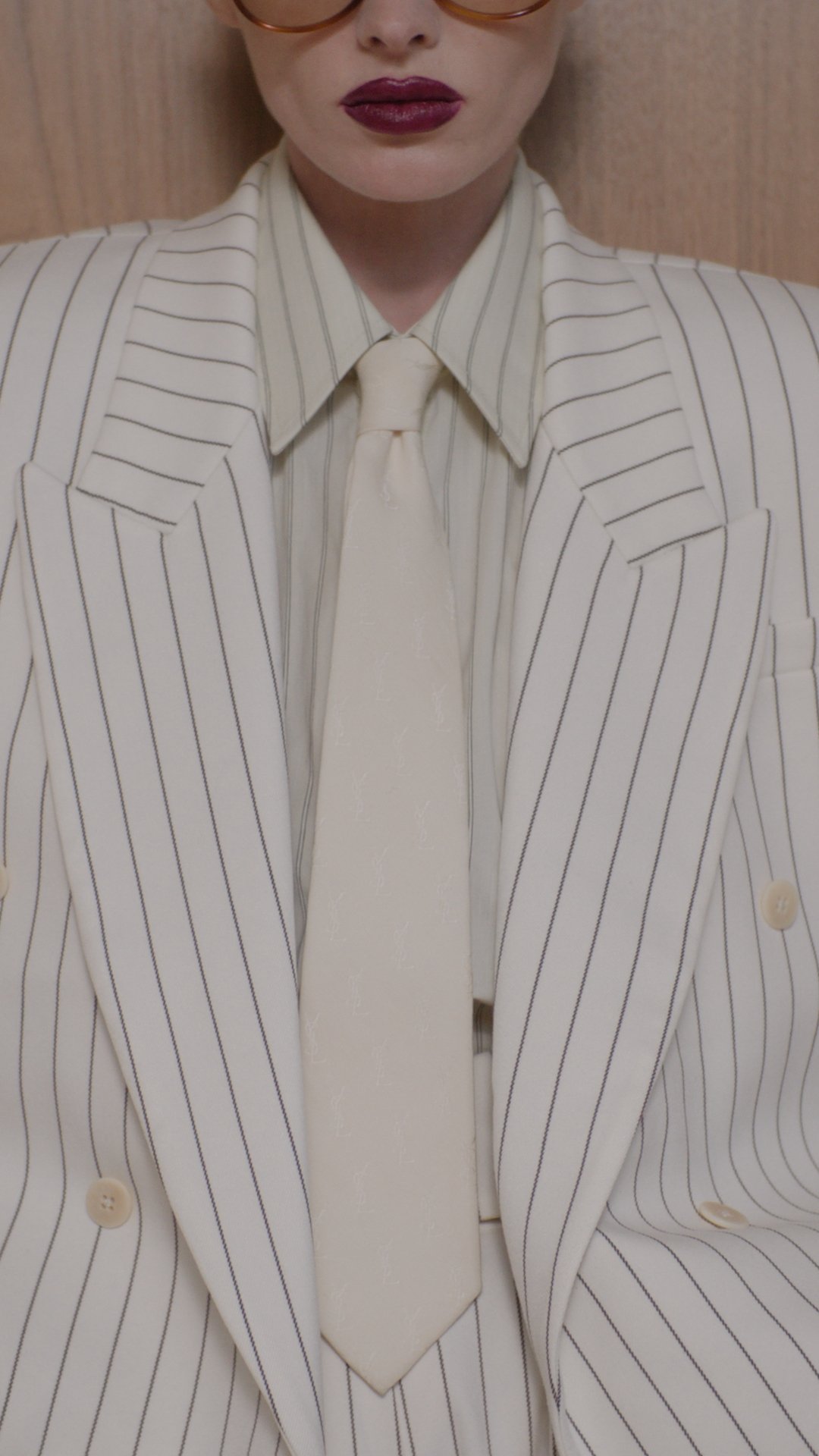 Close-up of a person wearing a beige pinstripe blazer with a matching shirt and tie, with dark lipstick and glasses.