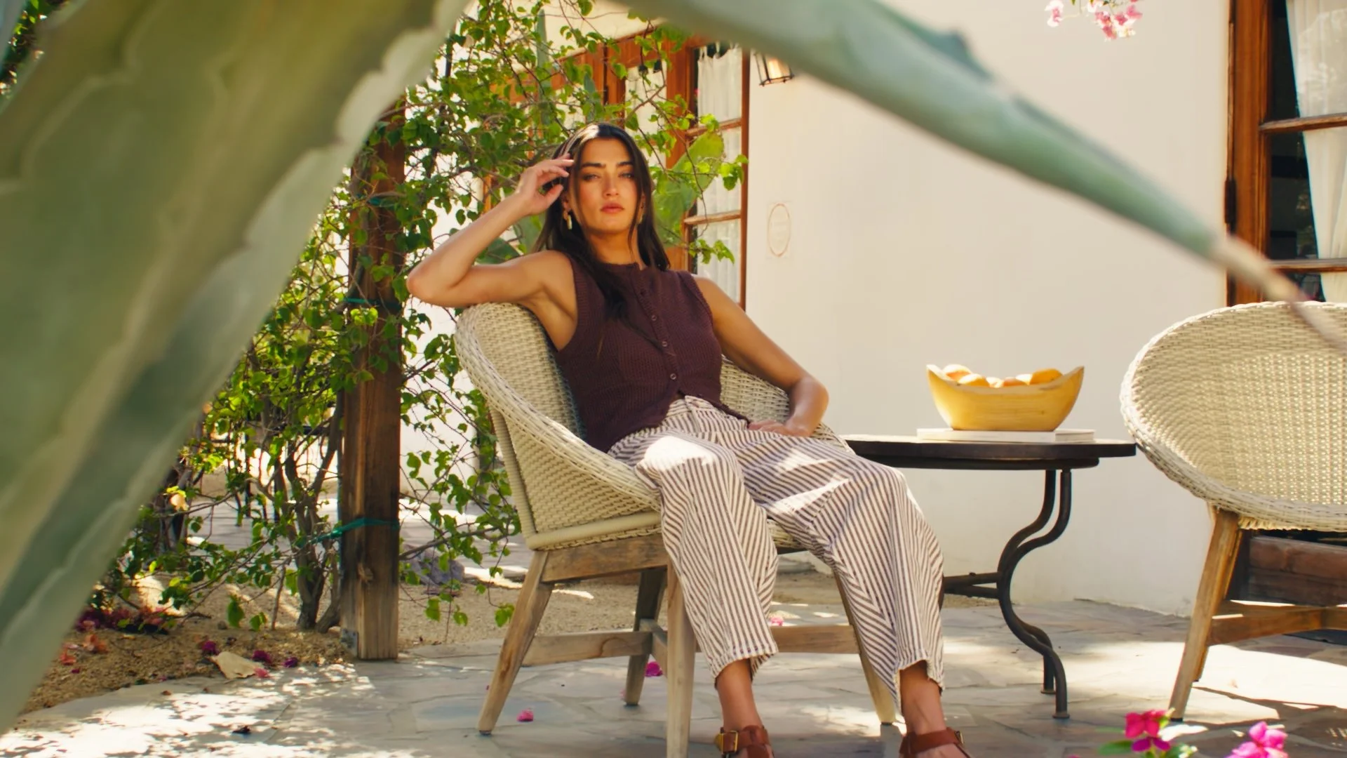 A woman sitting on a wicker chair outdoors, with greenery and a white wall in the background, wearing a sleeveless top and striped pants, next to a black table with a yellow bowl of fruit.