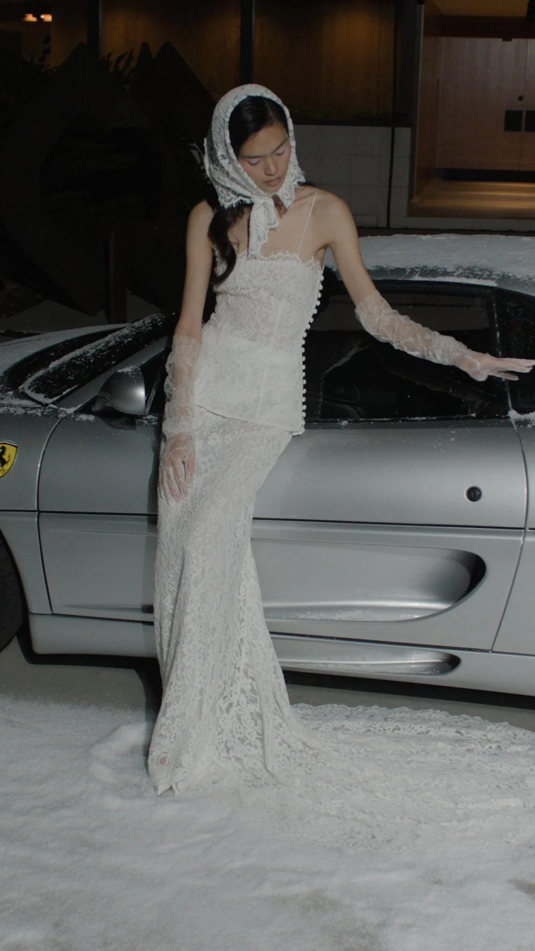 A woman in a white lace dress and lace gloves stands leaning on a silver Ferrari, with snow on the ground and snow dusted on the car.
