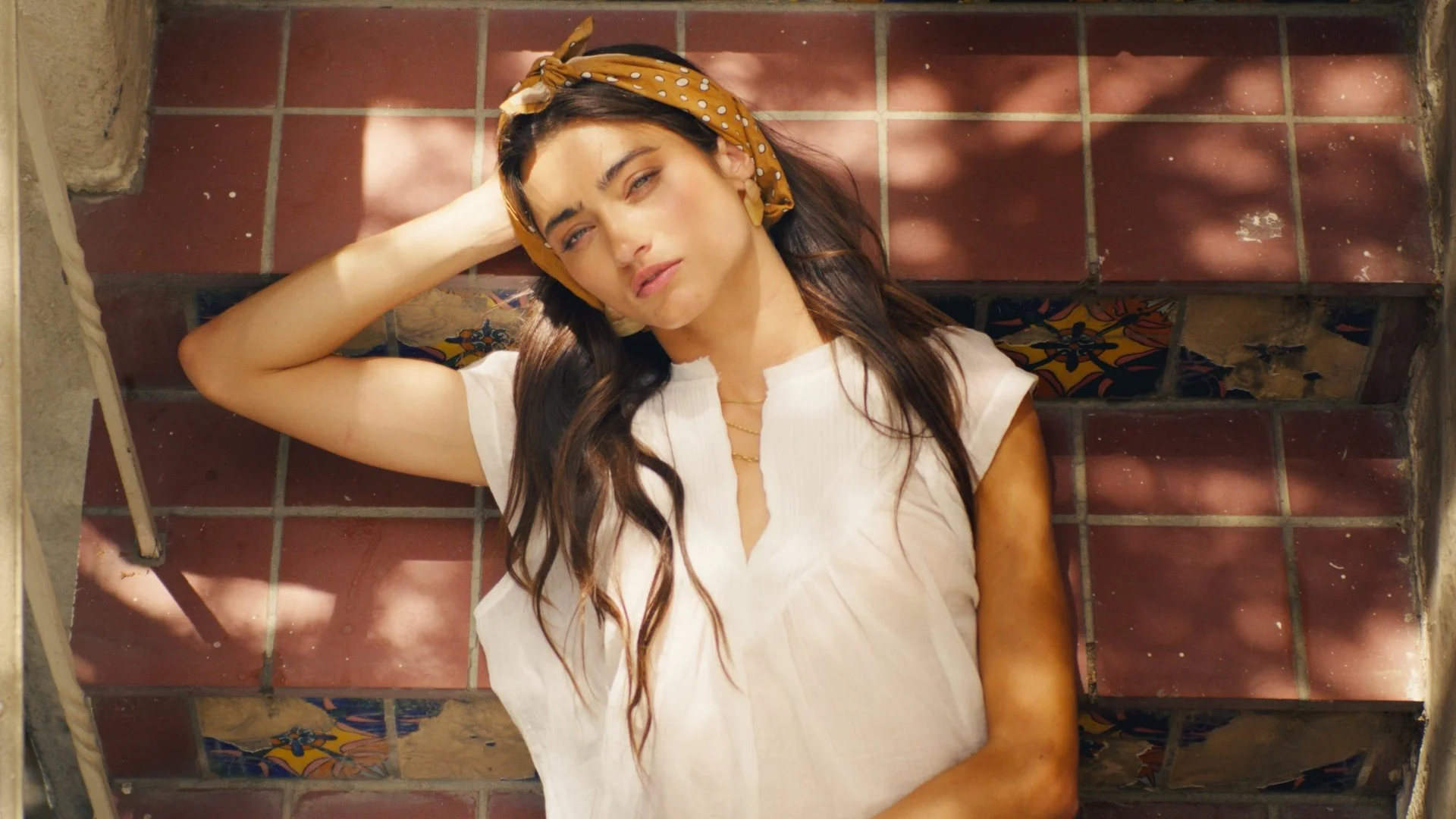 A woman lying on a tiled floor in the sun, wearing a beige top and a yellow polka dot headscarf.