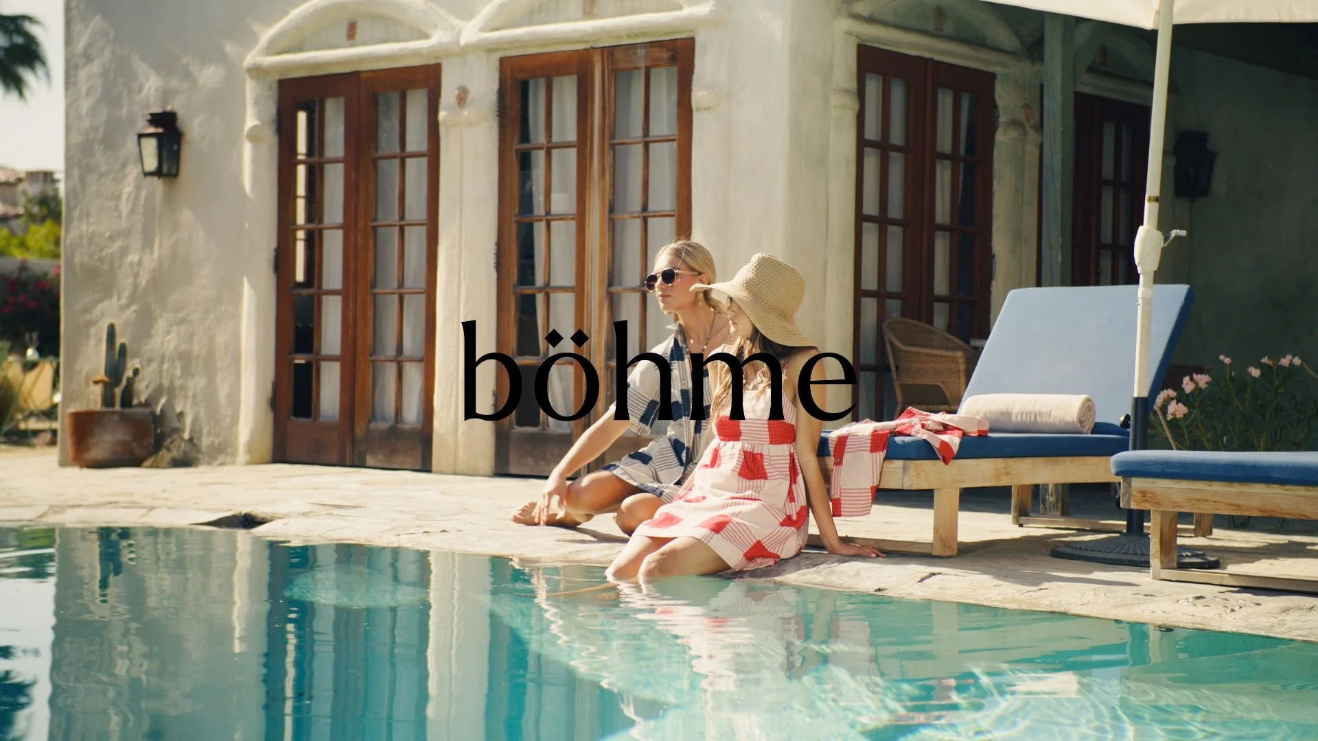Two women sitting by a swimming pool outside a house with large wooden doors and windows, one wearing sunglasses and a checkered shirt, the other wearing a sunhat and red and white checkered dress, with a lounge chair and a rolled towel nearby.