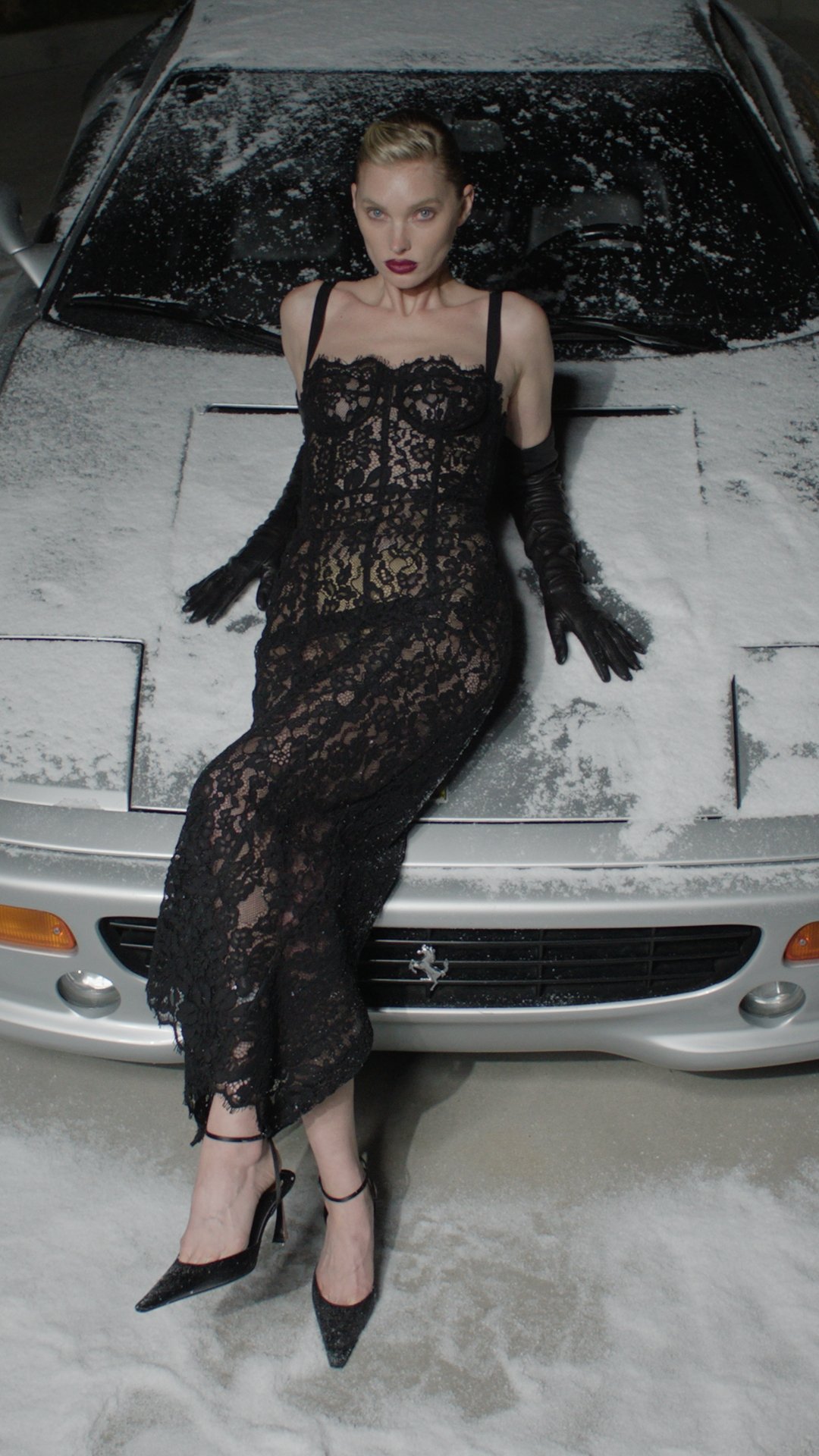 A woman in a black lace dress and high heels sitting on a snow-covered silver sports car.