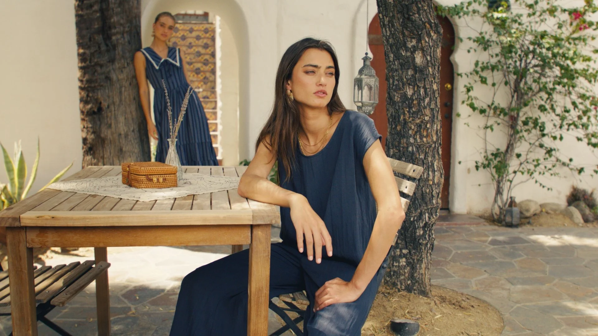 A woman with long dark hair wearing a dark blue dress, sitting outdoors at a wooden table under a tree, with a contemplative expression. In the background, another woman in a blue dress stands near a doorway, with potted plants and a white wall.