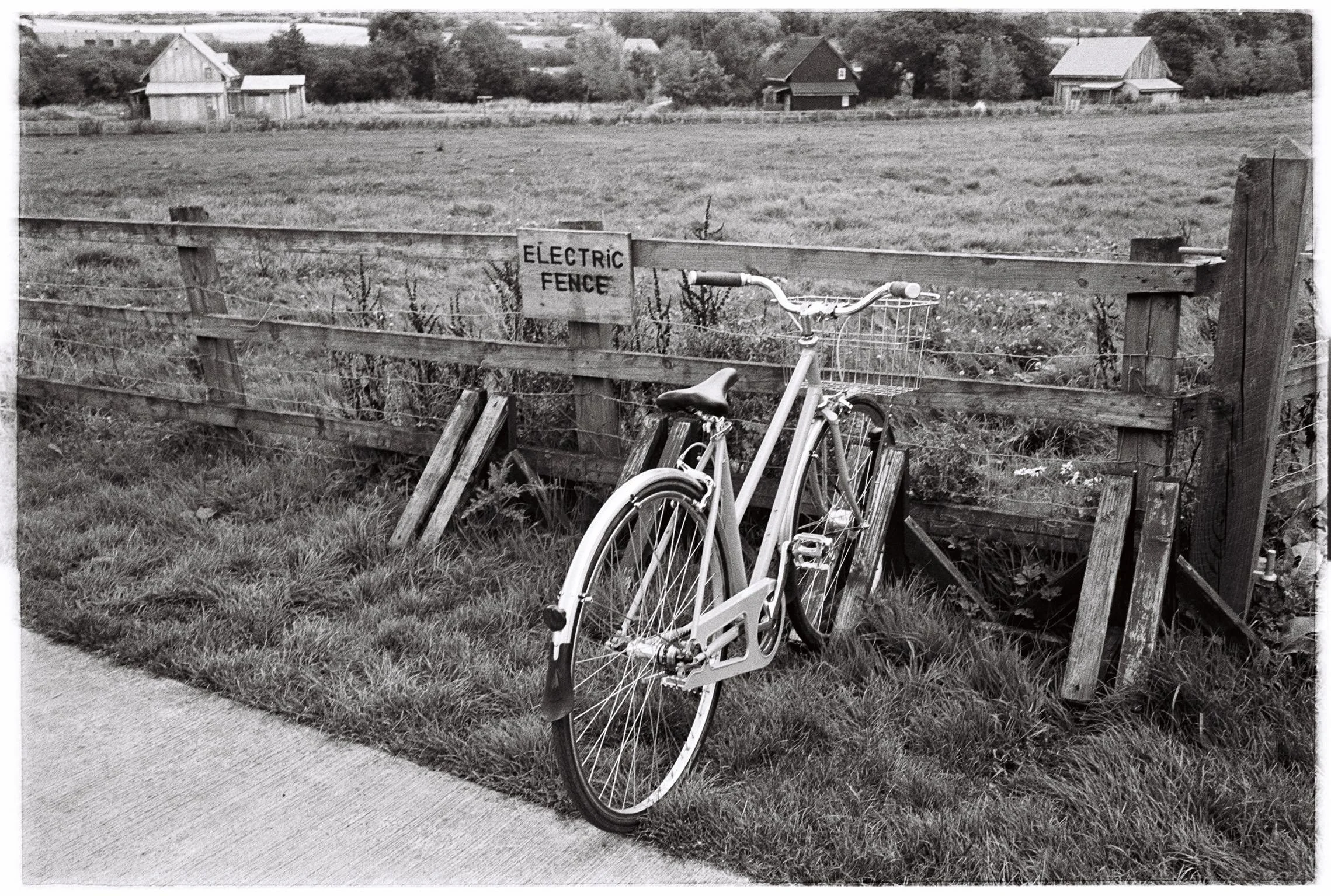 A black-and-white photograph of a bicycle leaning against a wooden fence in a rural setting. The fence has a sign that reads 'Electric Fence.' In the background, there are a few houses and trees, and an open grassy field.