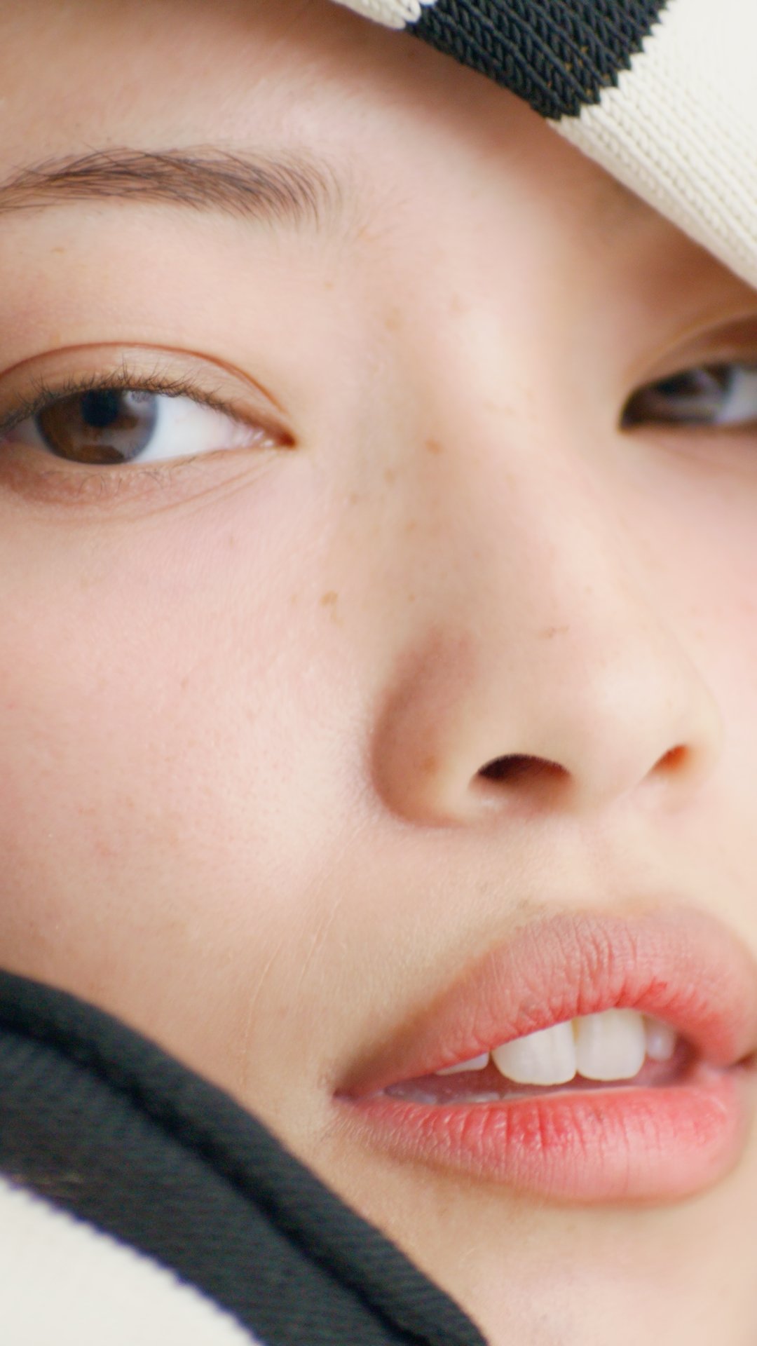 Close-up of a smiling woman's face, showing her eye, nose, and lips, with a black and white cap partially visible.