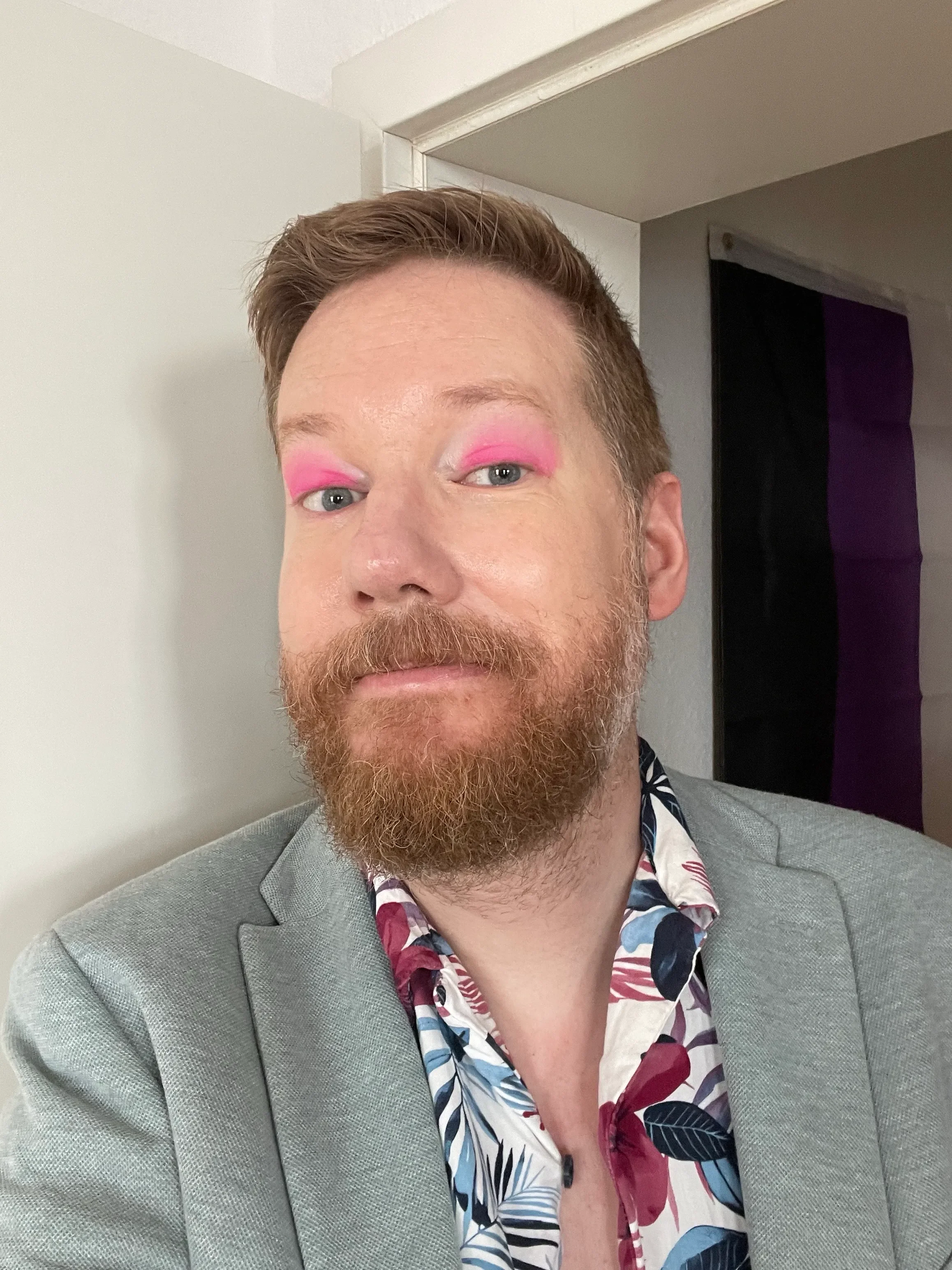 A selfie of Redfern Barrett wearing a magenta and deep blue floral shirt and light blue jacket. They have bright pink eyeshadow on and are standing in front of a non-bianry flag.