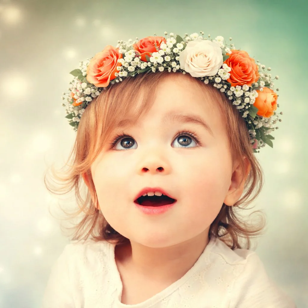 A photo of a very young child with strawberry blonde hair and hazel eyes looking upwards in amazement. The child is wearing a flower crown of orange and white silk roses and baby's breath.