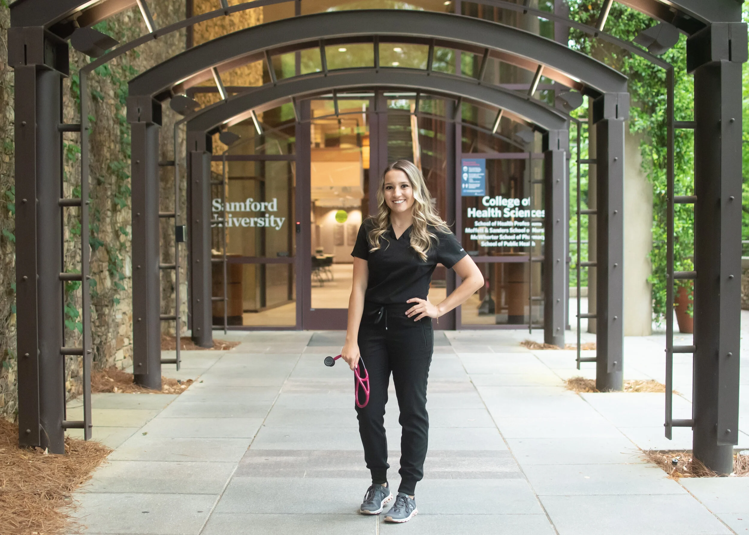 Young woman in black scrubs with stethoscope standing outside the entrance to the College of Health Sciences at Samford University, smiling.