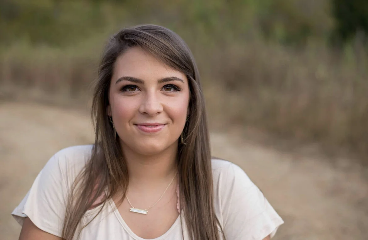 A young woman with long brown hair smiling outdoors, wearing a white top and a gold necklace.