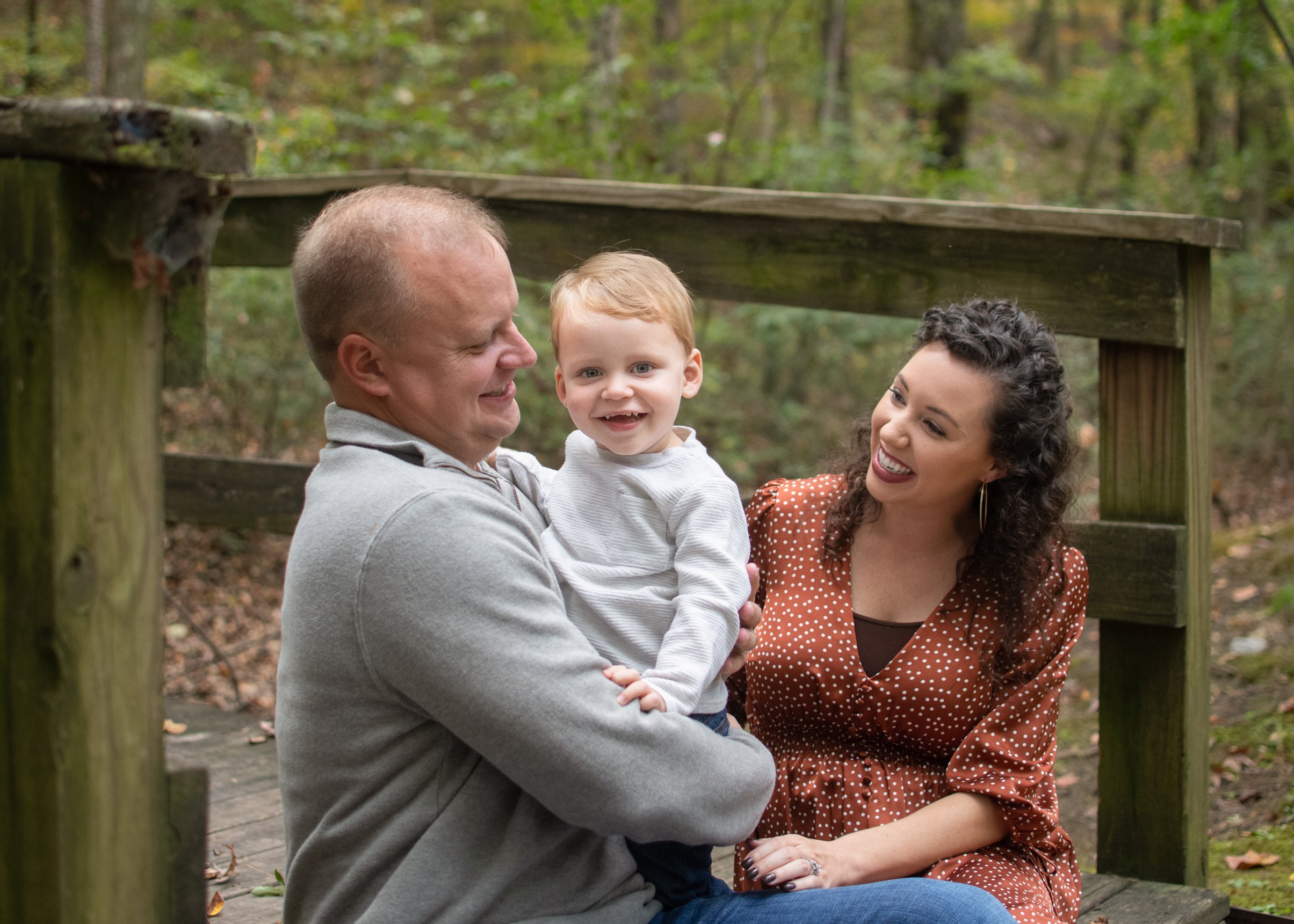A family of three enjoying time outdoors in a wooded area. The father is holding a young boy, and the mother is sitting next to them, all smiling and looking at each other.