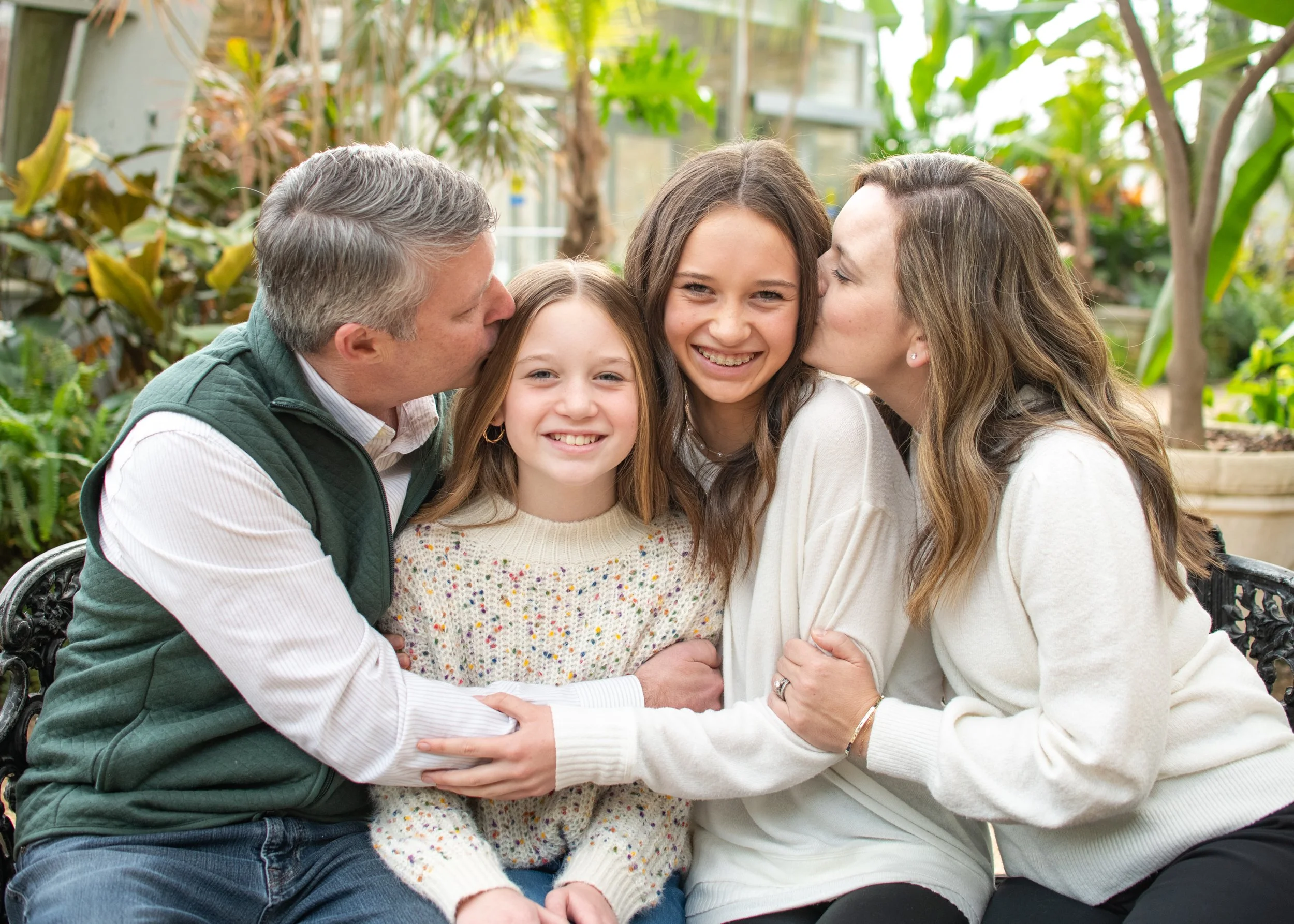 Family of four sitting on a bench in a greenhouse; parents kissing their two daughters on the cheeks, all smiling.