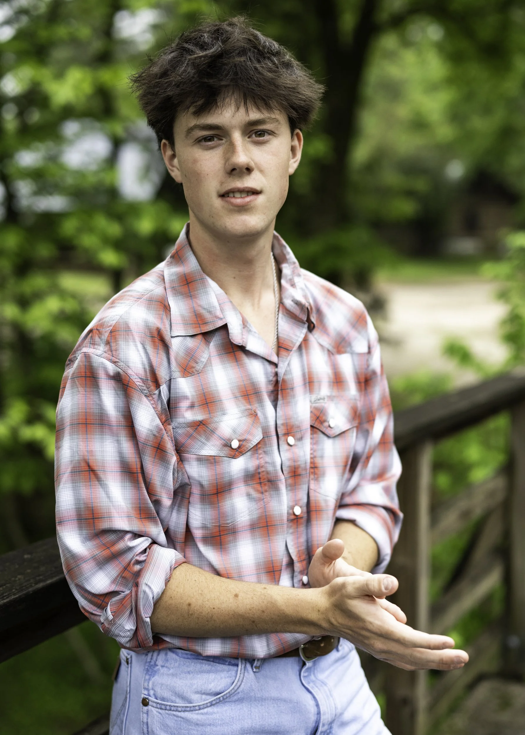 A young man with messy brown hair standing outdoors on a wooden bridge, wearing a red and white checkered shirt with rolled-up sleeves and light blue jeans, with greenery and trees in the background.
