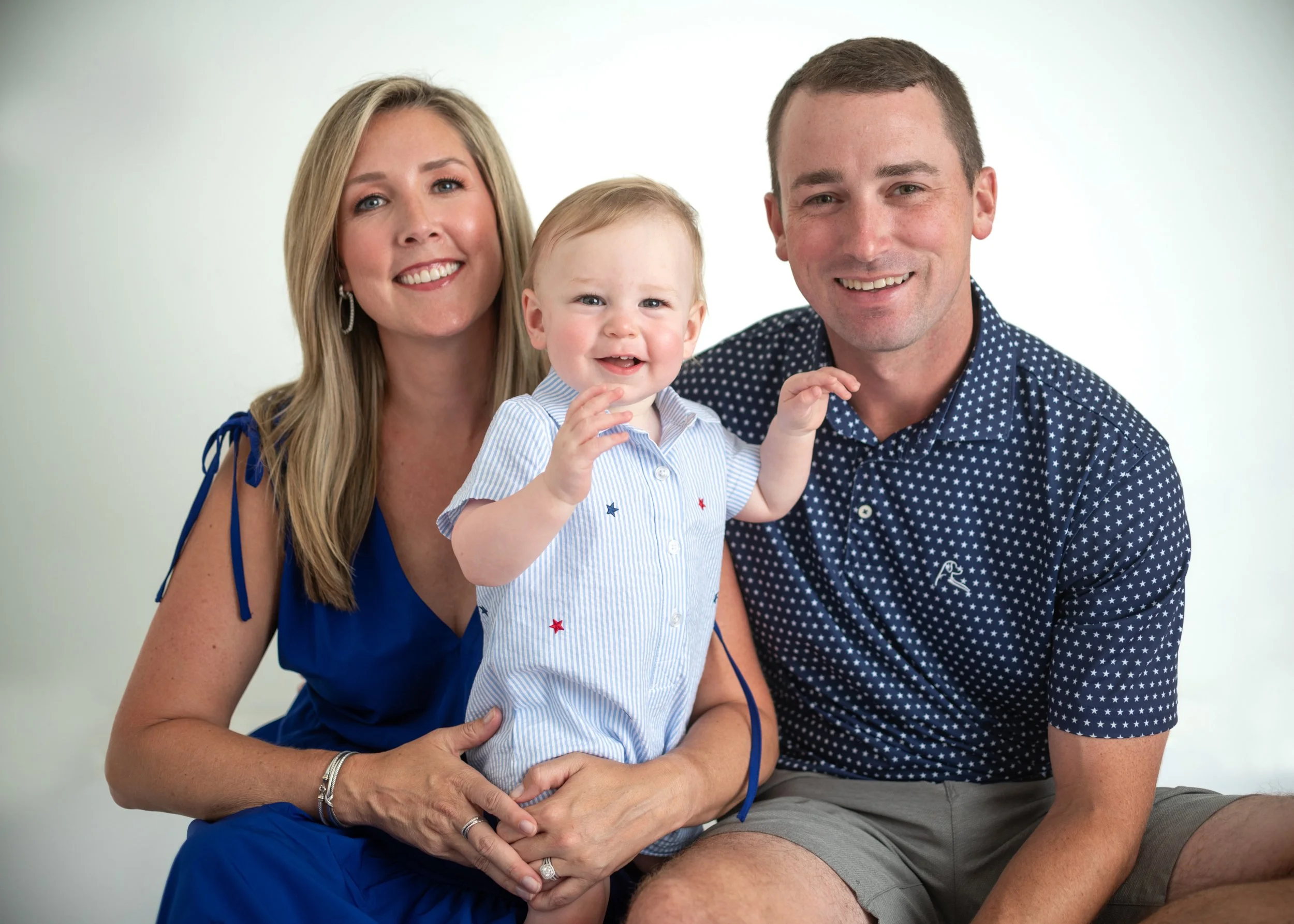 A smiling woman with blonde hair in a blue dress, a young boy in a light blue striped shirt with stars, and a smiling man with short brown hair in a navy blue polka dot shirt, sitting together against a white background.