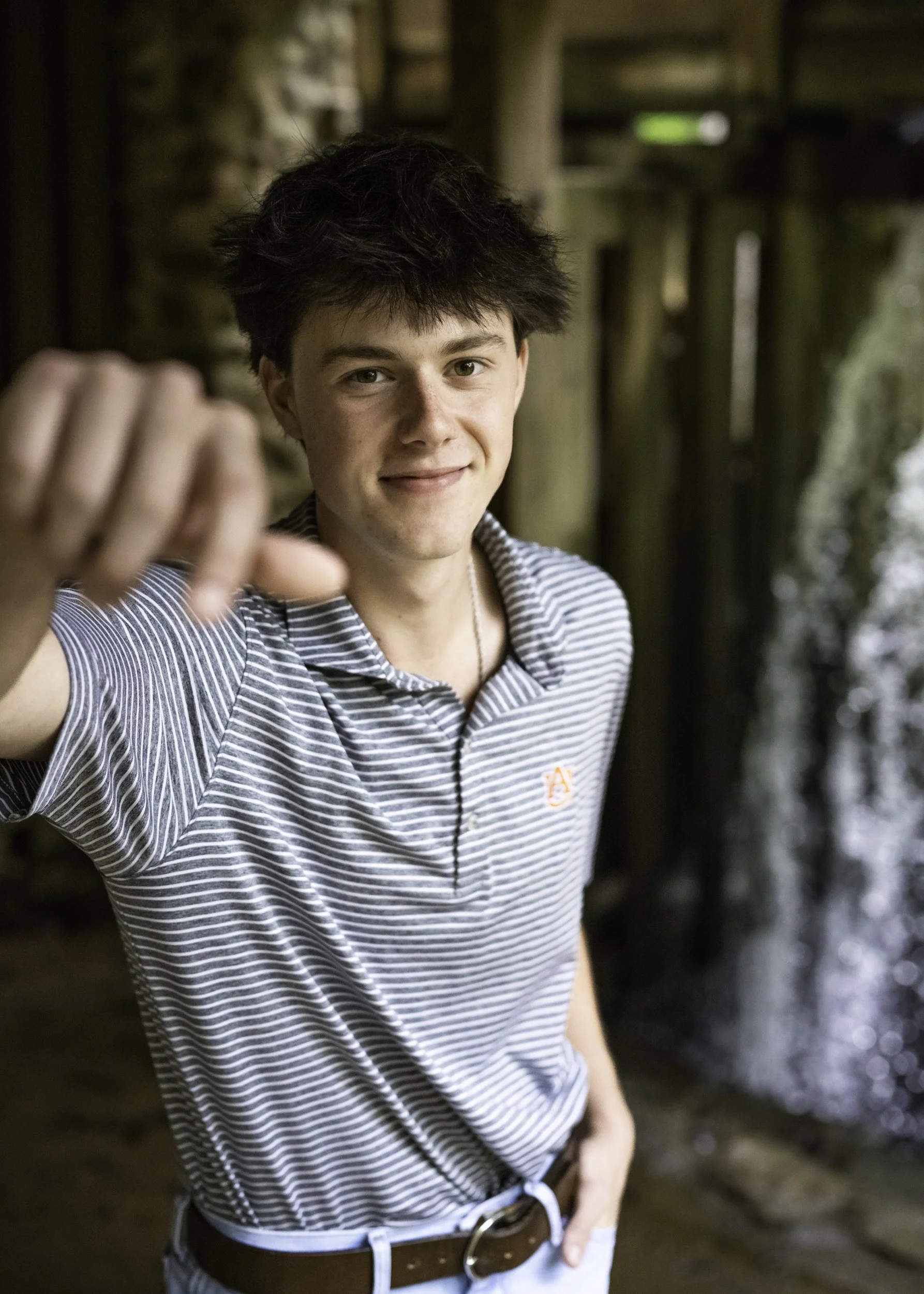 A young man with dark, messy hair smiling and reaching out towards the camera. He is wearing a striped polo shirt and standing outdoors near a waterfall.
